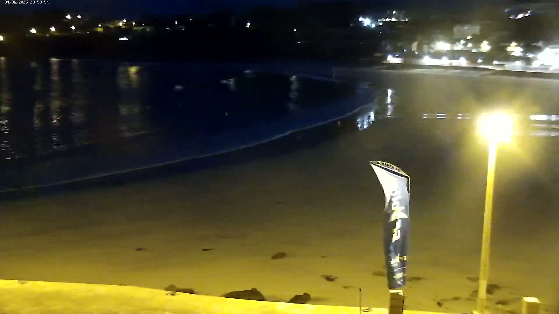 A dark, sandy beach at night is gently lapped by small waves under a clear, moonless sky, illuminated by a single lamppost.