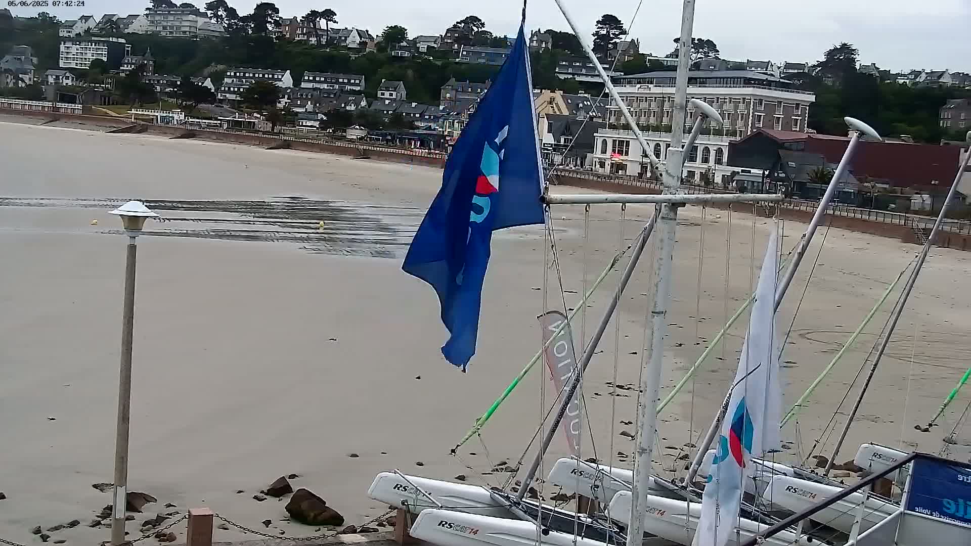 A sandy beach with several small sailboats beached on it, a flagpole with a blue flag, and a coastal town visible in the background under an overcast sky.