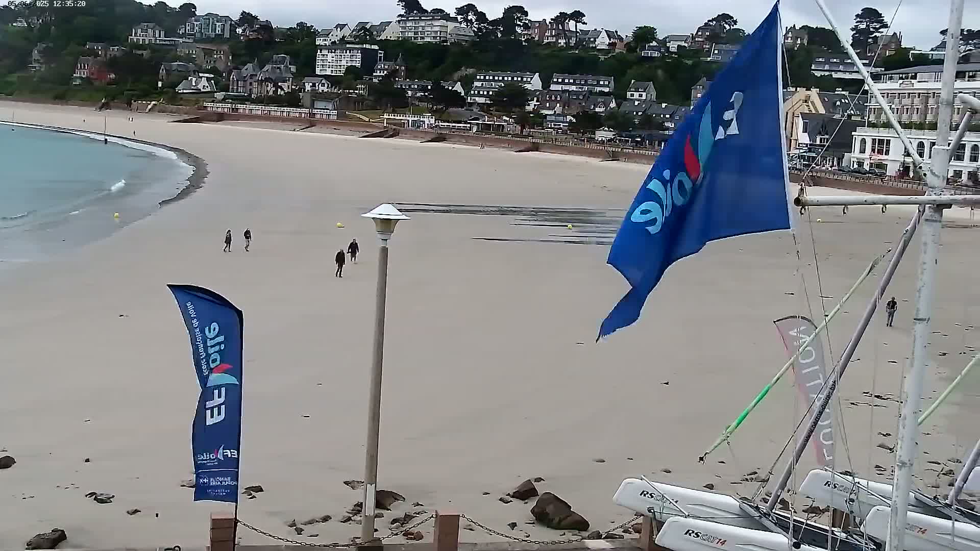 A sandy beach with a few people walking on it, boats grounded on the shore, and a town on a hill in the background under an overcast sky.