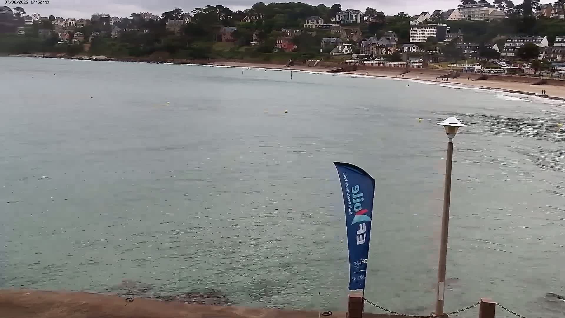 A calm, overcast day at a beach with a town of houses on a hill overlooking the water, a sandy beach, and a flagpole with a flag.