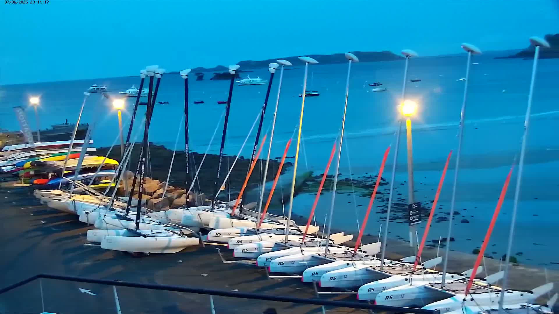 Numerous small sailboats are docked along a rocky shore at twilight under a clear, dark blue sky.