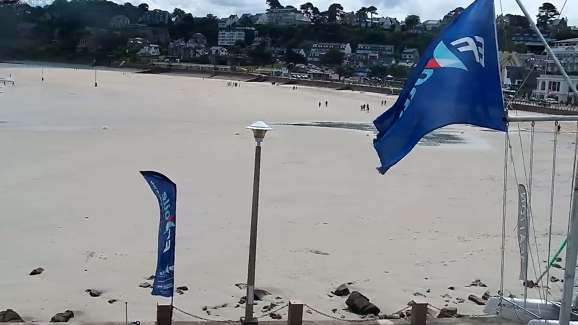 A mostly empty sandy beach with a few people and buildings in the background on a cloudy day, with two flags waving in the wind.