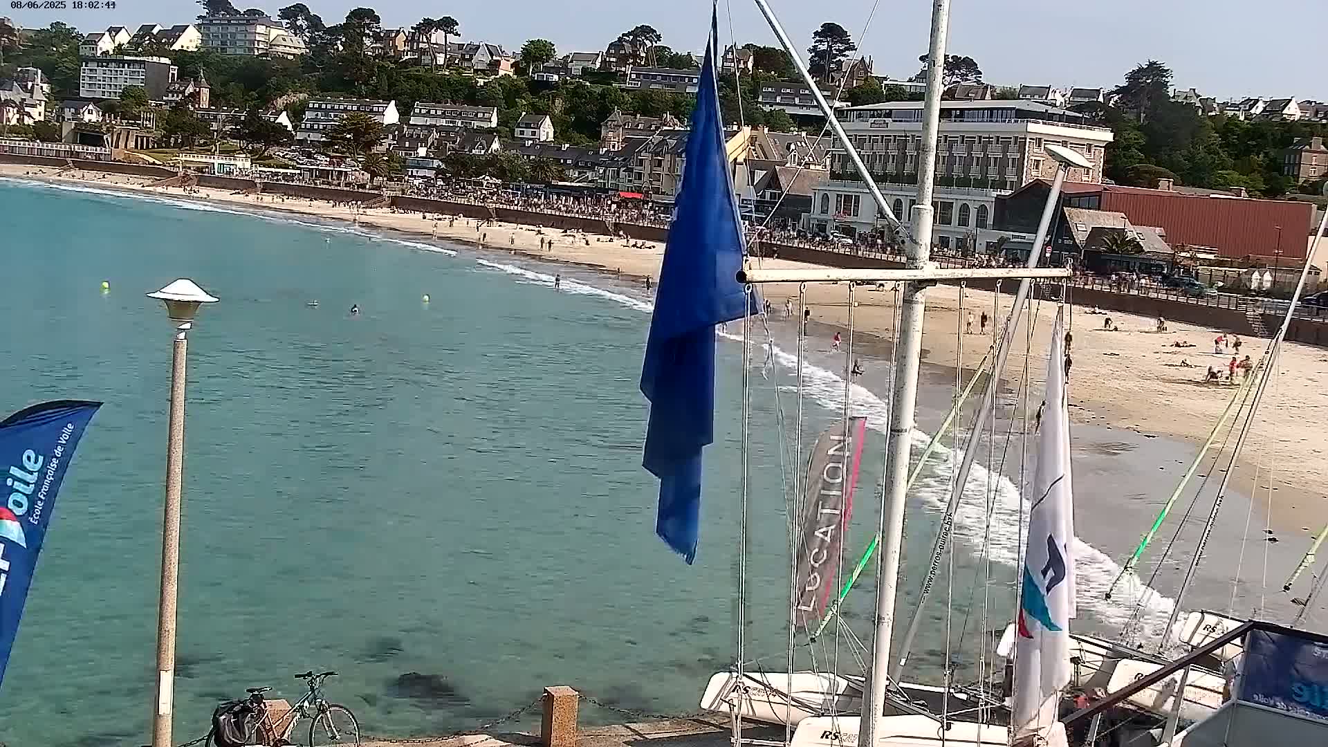 A sandy beach with many people on it is next to a calm, teal-colored ocean; several sailboats are docked nearby, and a town is visible in the background on a sunny day.