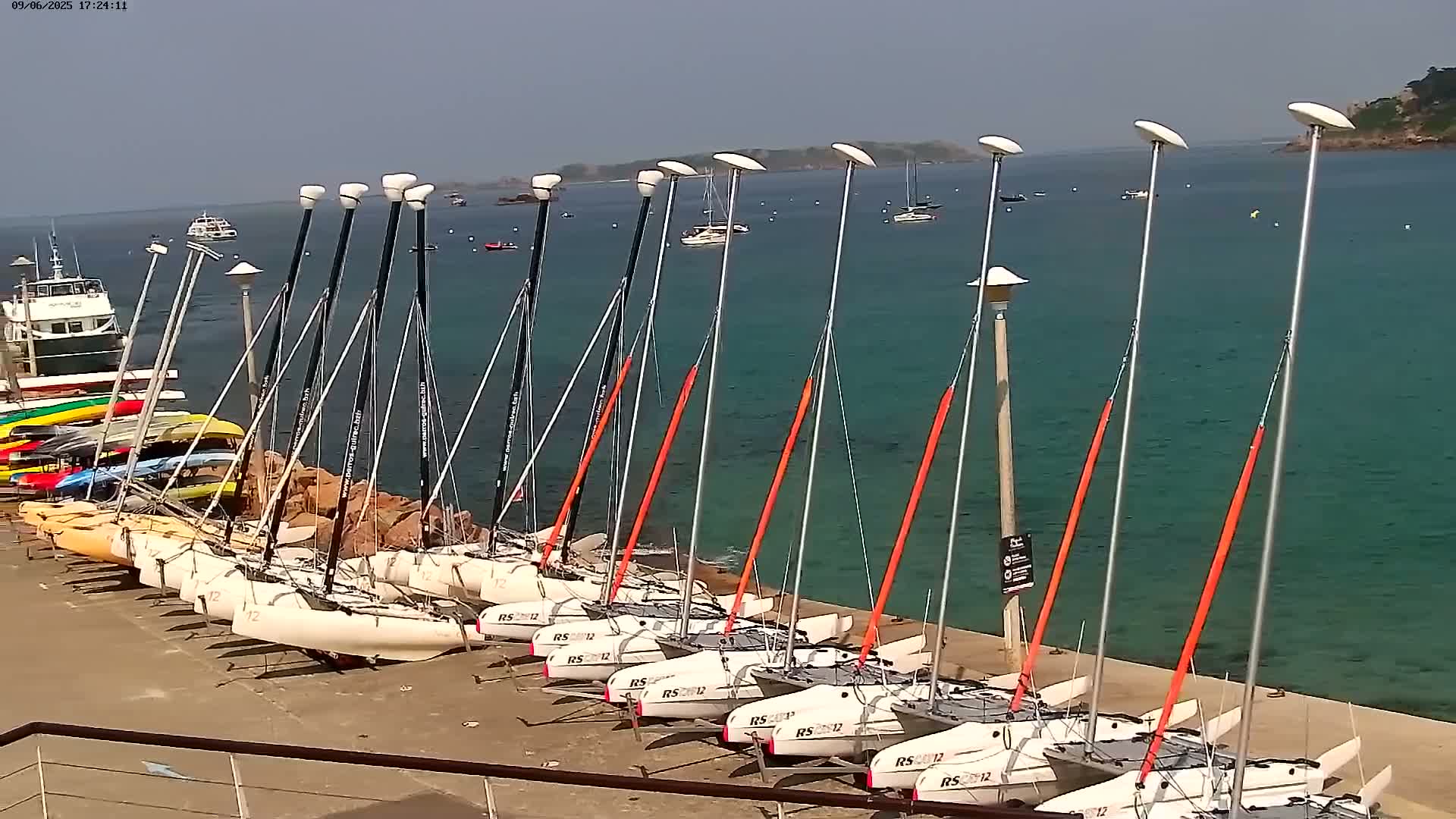 Numerous small sailboats are docked along a shoreline on a sunny day with calm, teal water.