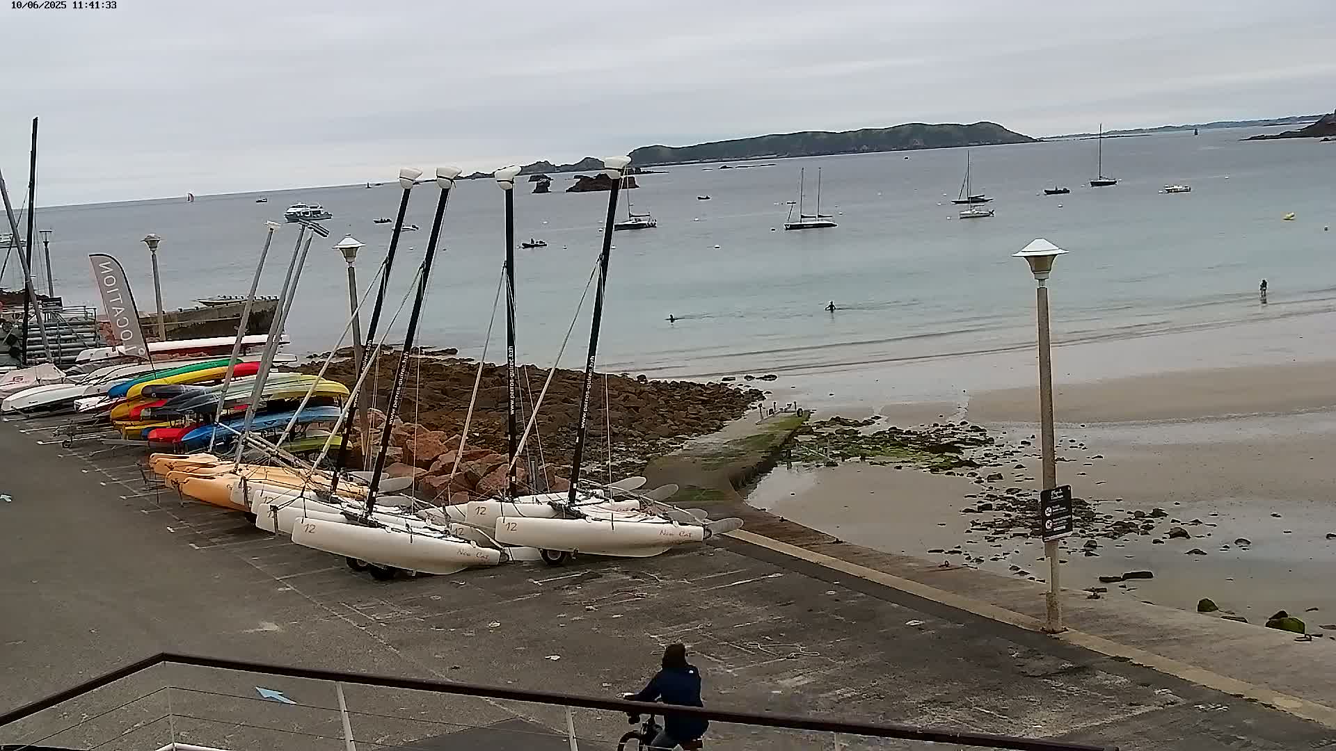 Several sailboats and catamarans are parked on a paved area next to a sandy beach, with a calm sea and overcast sky in the background.