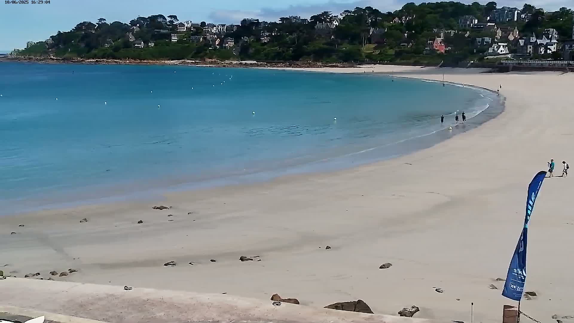 A wide sandy beach curves around a calm, turquoise bay under a clear sky, with a few people wading in the shallows and walking along the shore.