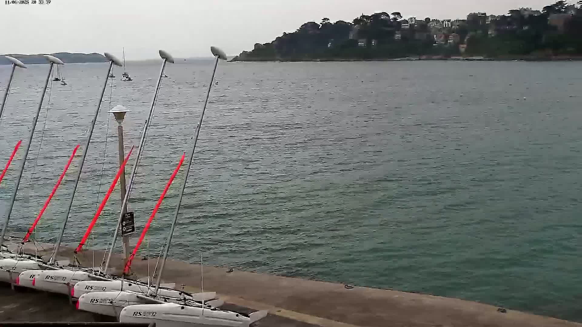 Several small sailboats are moored at a dock next to calm, dark-green water under an overcast sky.
