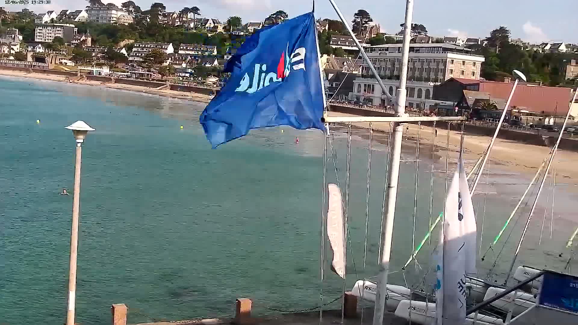 A calm, sunny day at a beach with a town in the background, featuring a blue flag, several sailboats docked in shallow water, and a person swimming in the ocean.