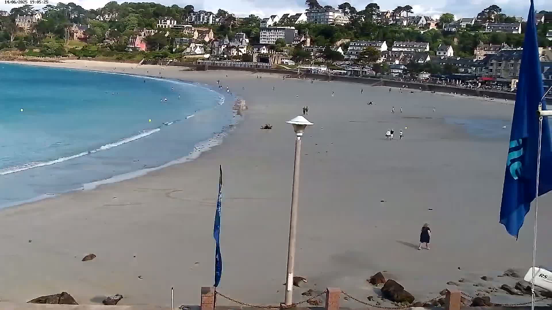 A sandy beach with a few people and gentle waves laps a shoreline in front of a town of buildings on a hillside under a partly cloudy sky.