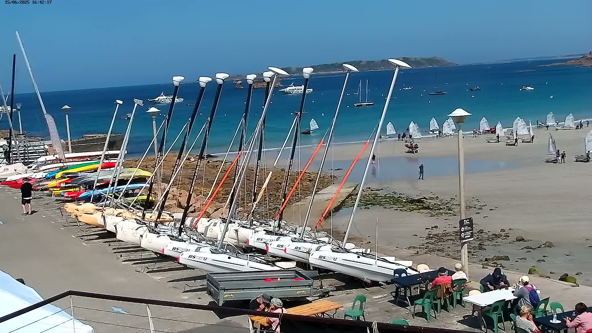 A sunny day shows numerous sailboats docked at a rocky shore near a sandy beach with additional sailboats on the water and beach.
