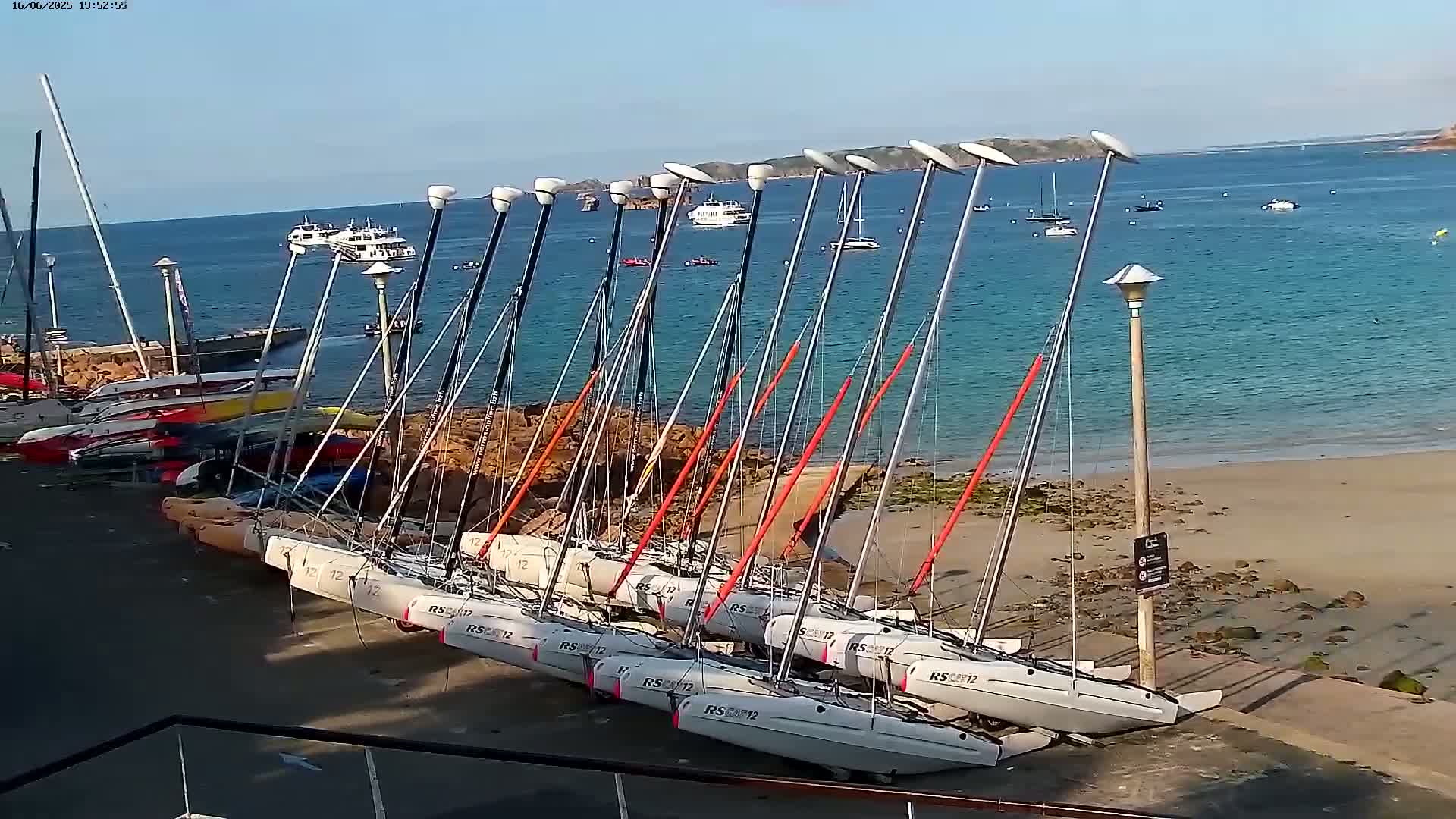 Numerous small sailboats are beached on a sandy shore near calm, blue water under a clear sky.