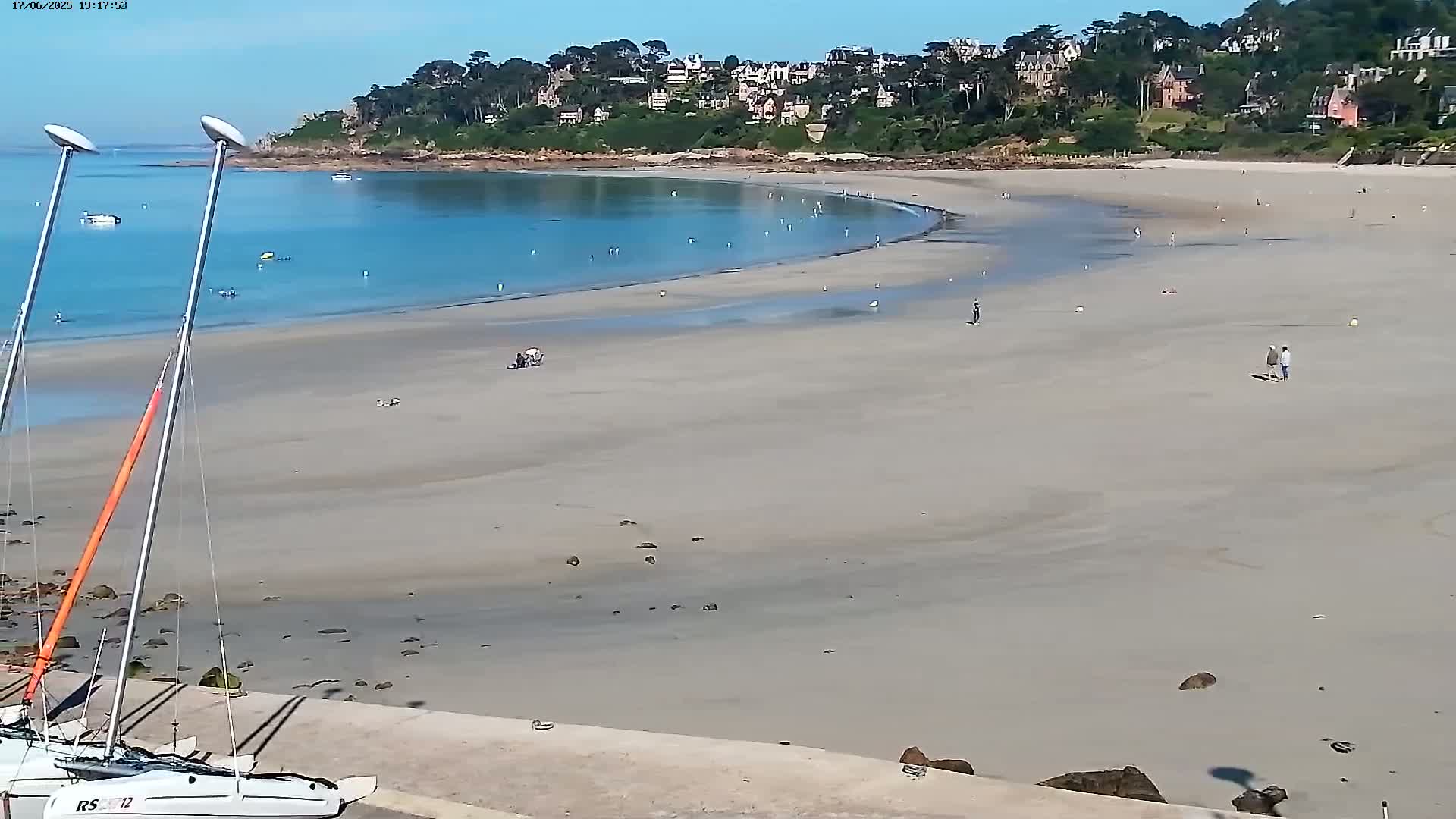 A wide sandy beach with shallow turquoise water curves around a headland where houses are built, under a clear sunny sky.