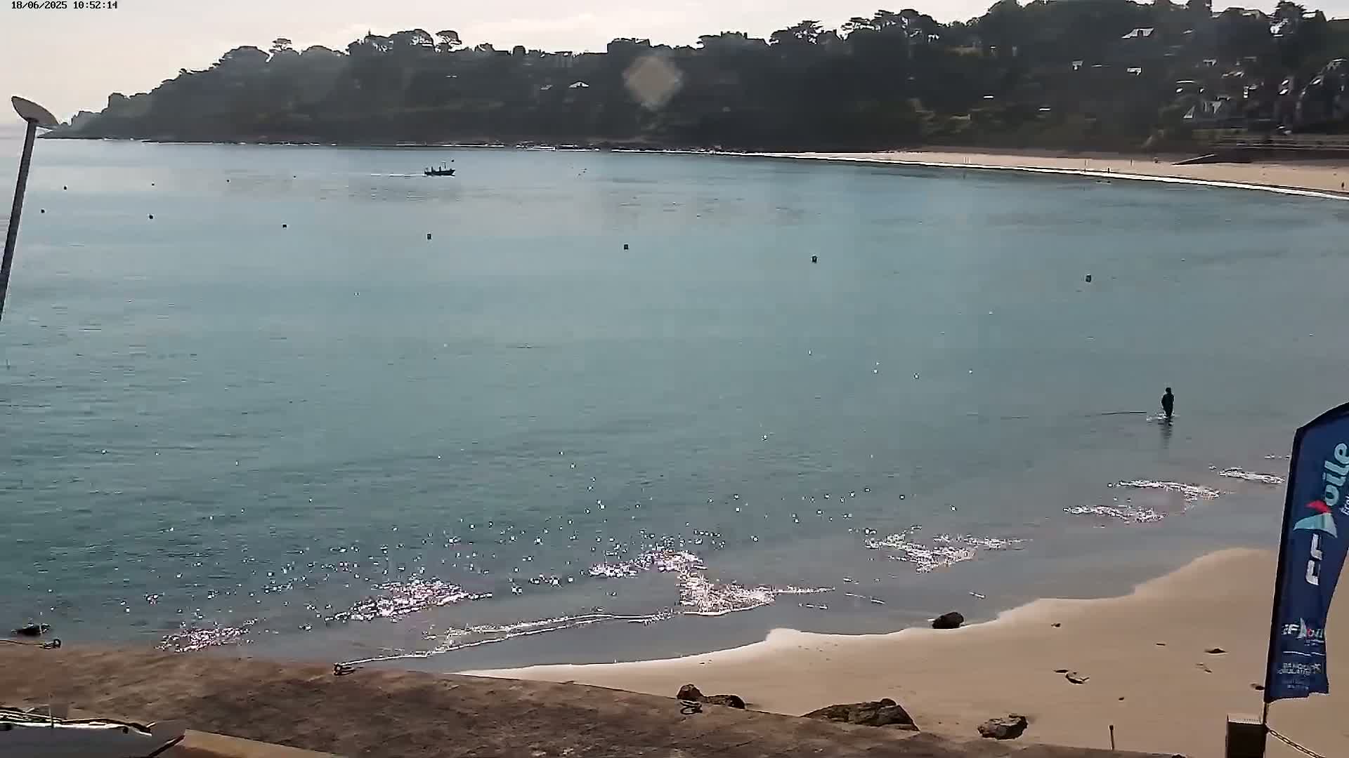 A calm, light-blue body of water laps a sandy beach, with a small boat in the distance and a person wading in the shallows, under an overcast sky.