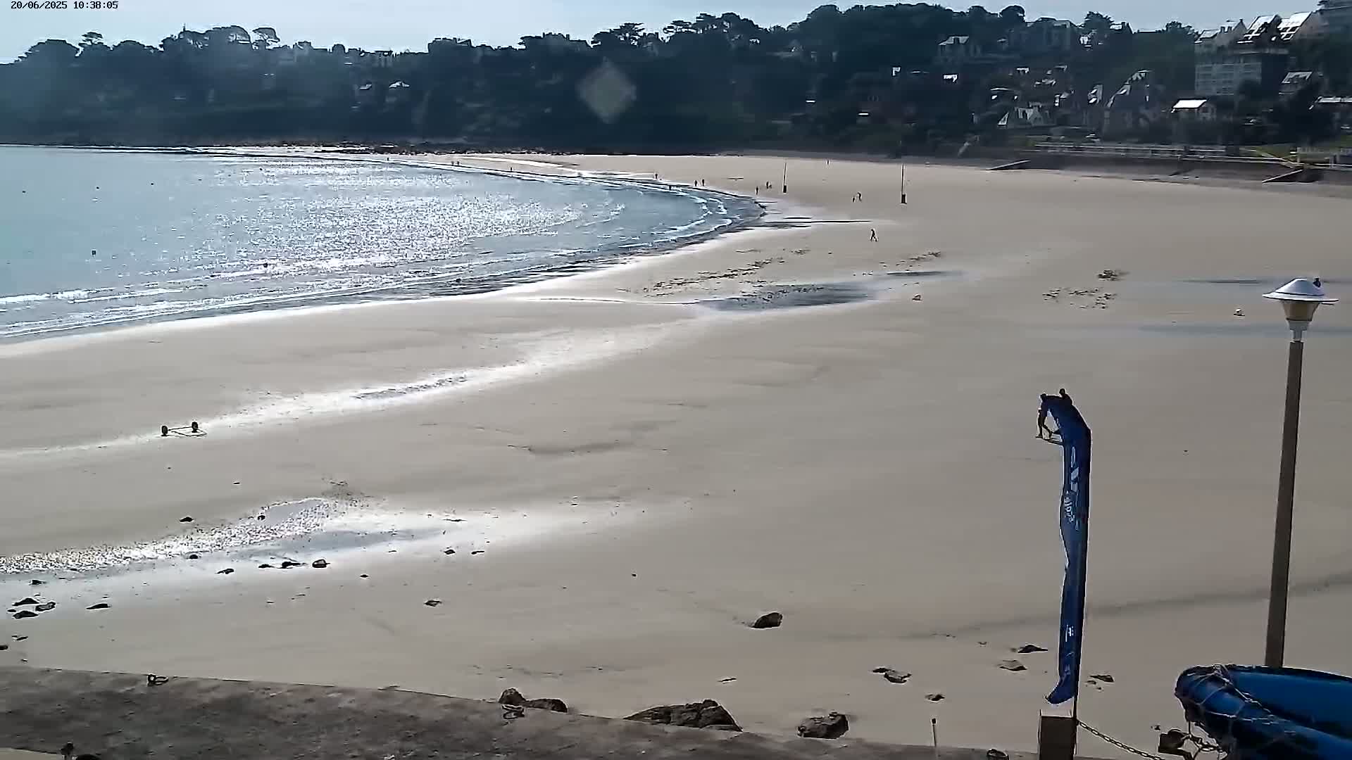 A sandy beach with gentle waves laps the shore under a mostly sunny sky, with a few people scattered on the sand and in the water, backed by a line of buildings and trees.