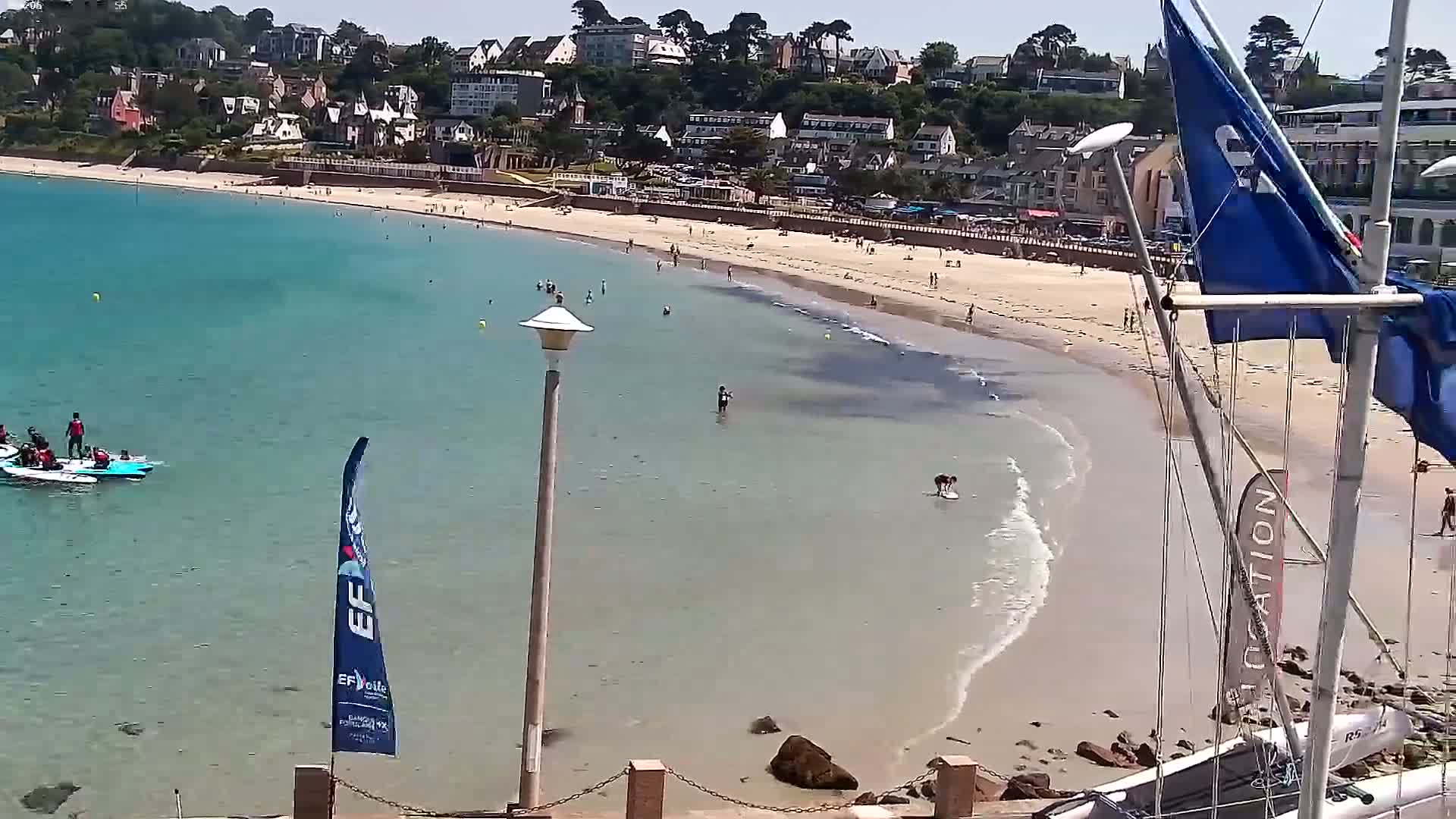 A sandy beach with clear, shallow turquoise water is partially occupied by swimmers and a small group in boats, all under a sunny sky, next to a coastal town with buildings on a hillside.