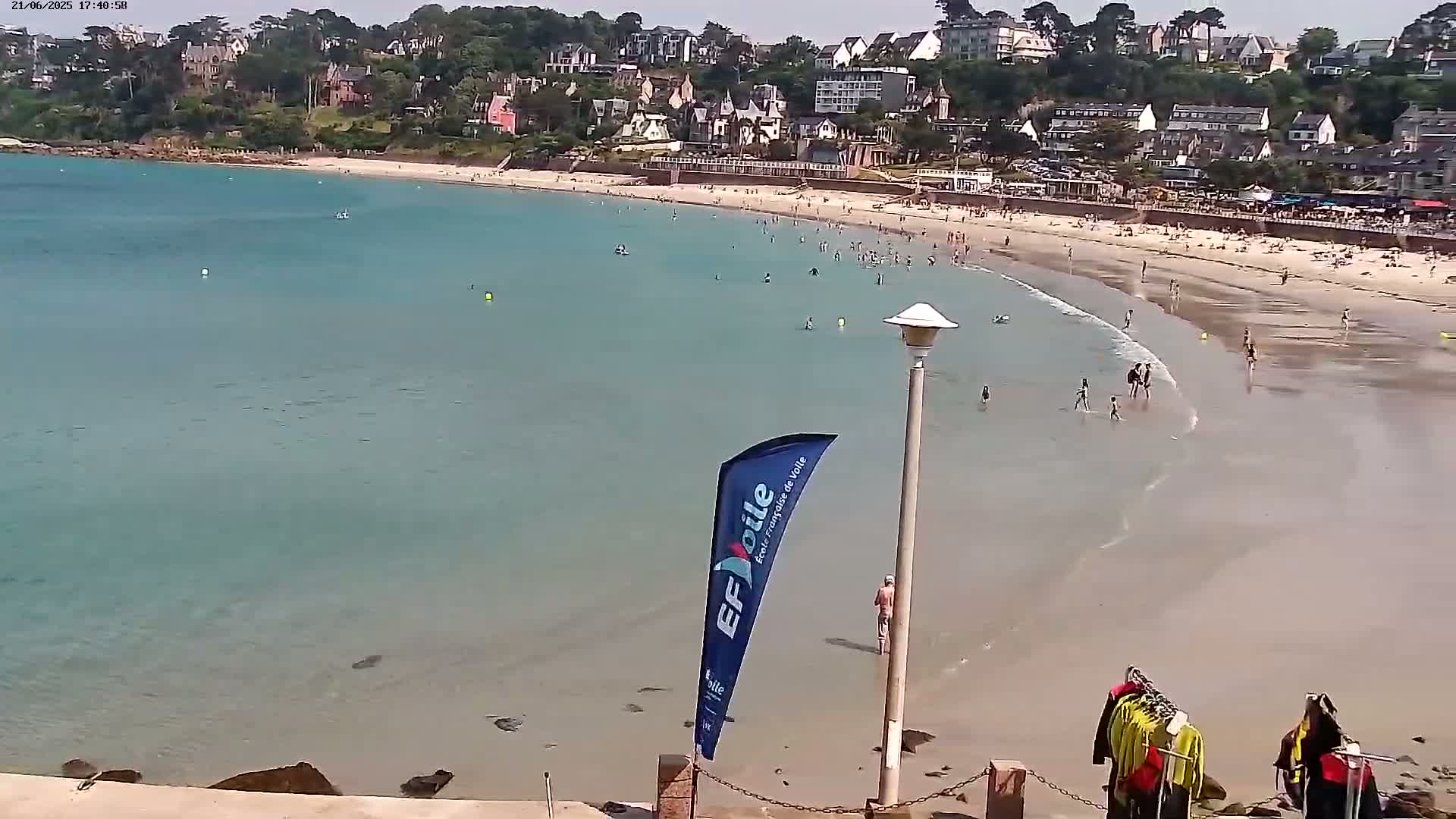 A sandy beach with calm, shallow, turquoise water is populated by swimmers and sunbathers, backed by a hillside town on a sunny day.
