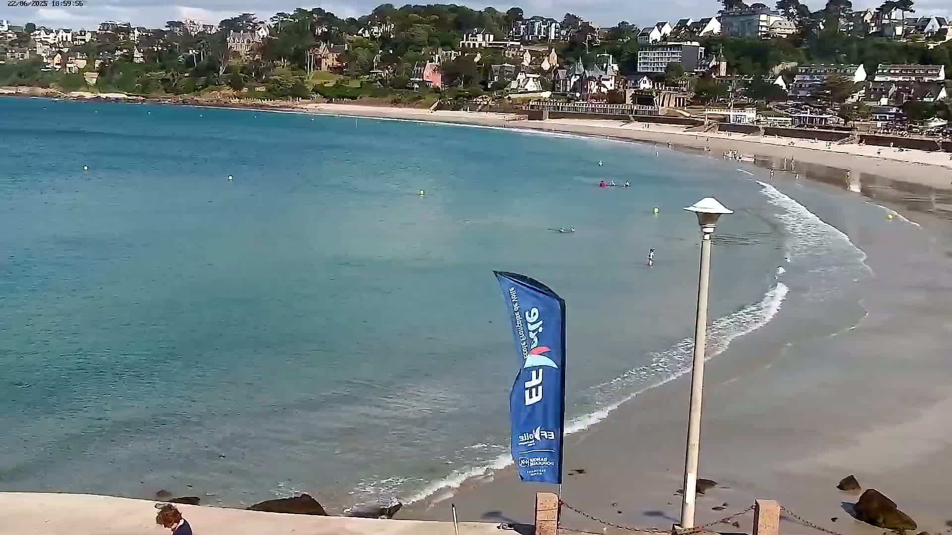 A sandy beach with calm, light-blue water curves around a shoreline dotted with buildings, under a mostly sunny sky.