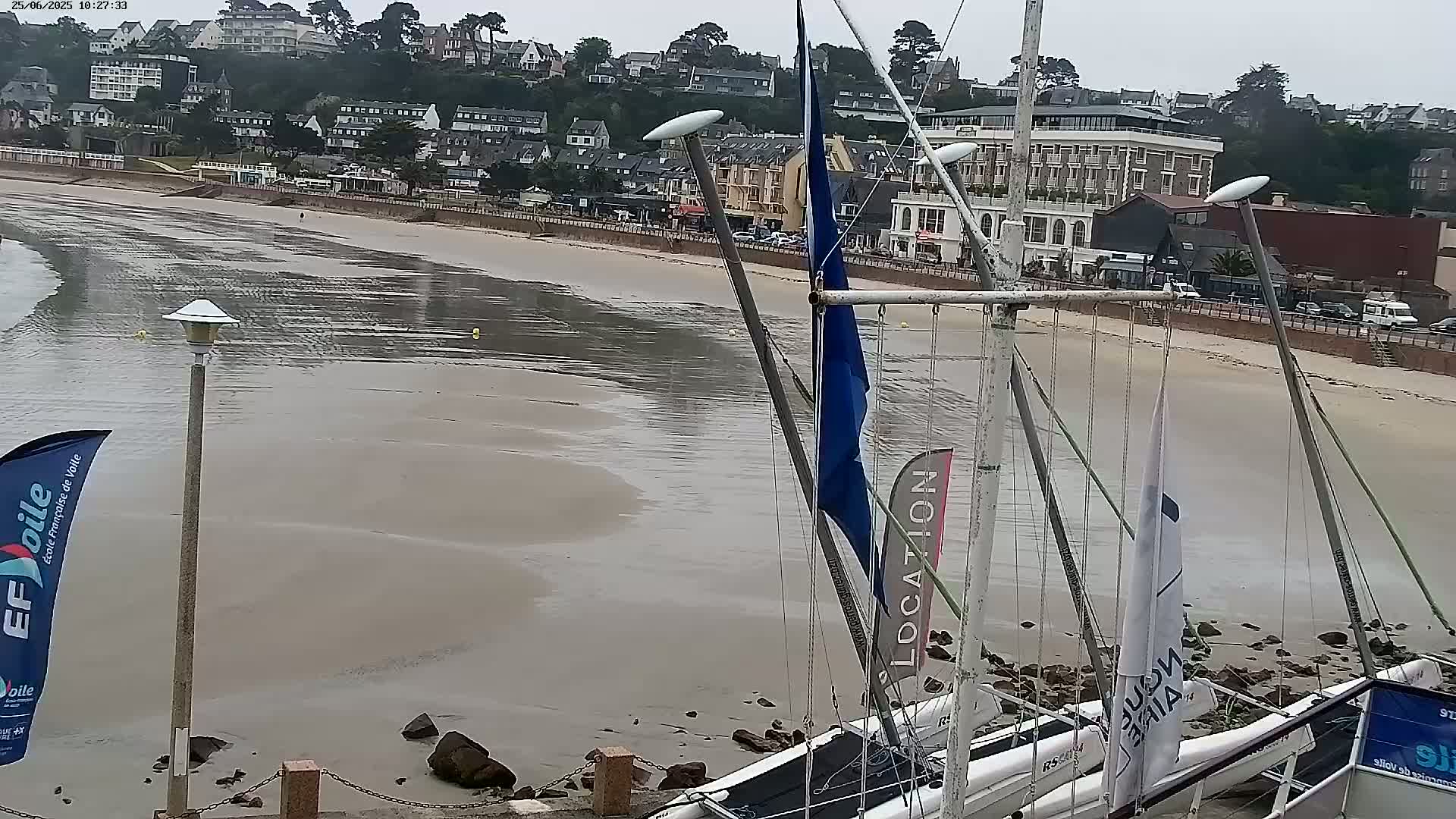 A mostly receding sandy beach with several sailboats beached on rocks at low tide near a seaside town under an overcast sky.
