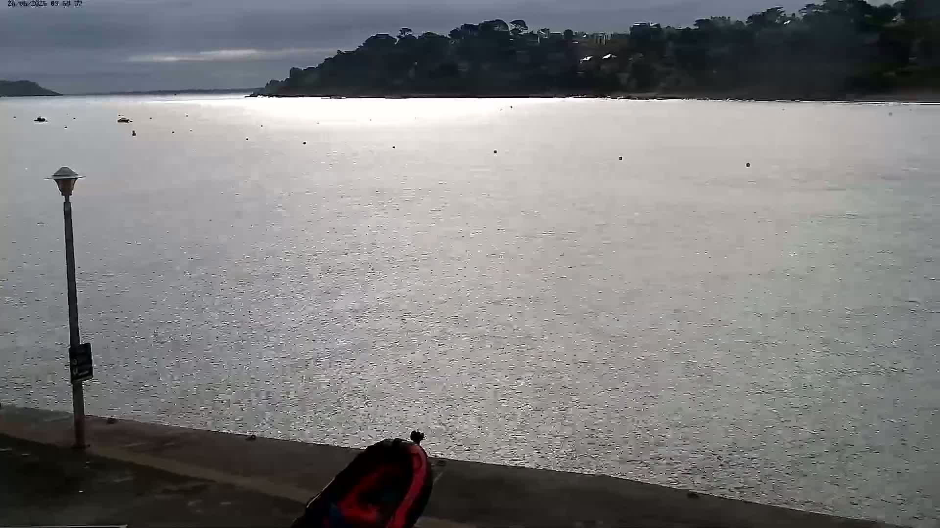 A calm body of water stretches out under an overcast sky, with a distant shoreline lined with trees and buildings visible.