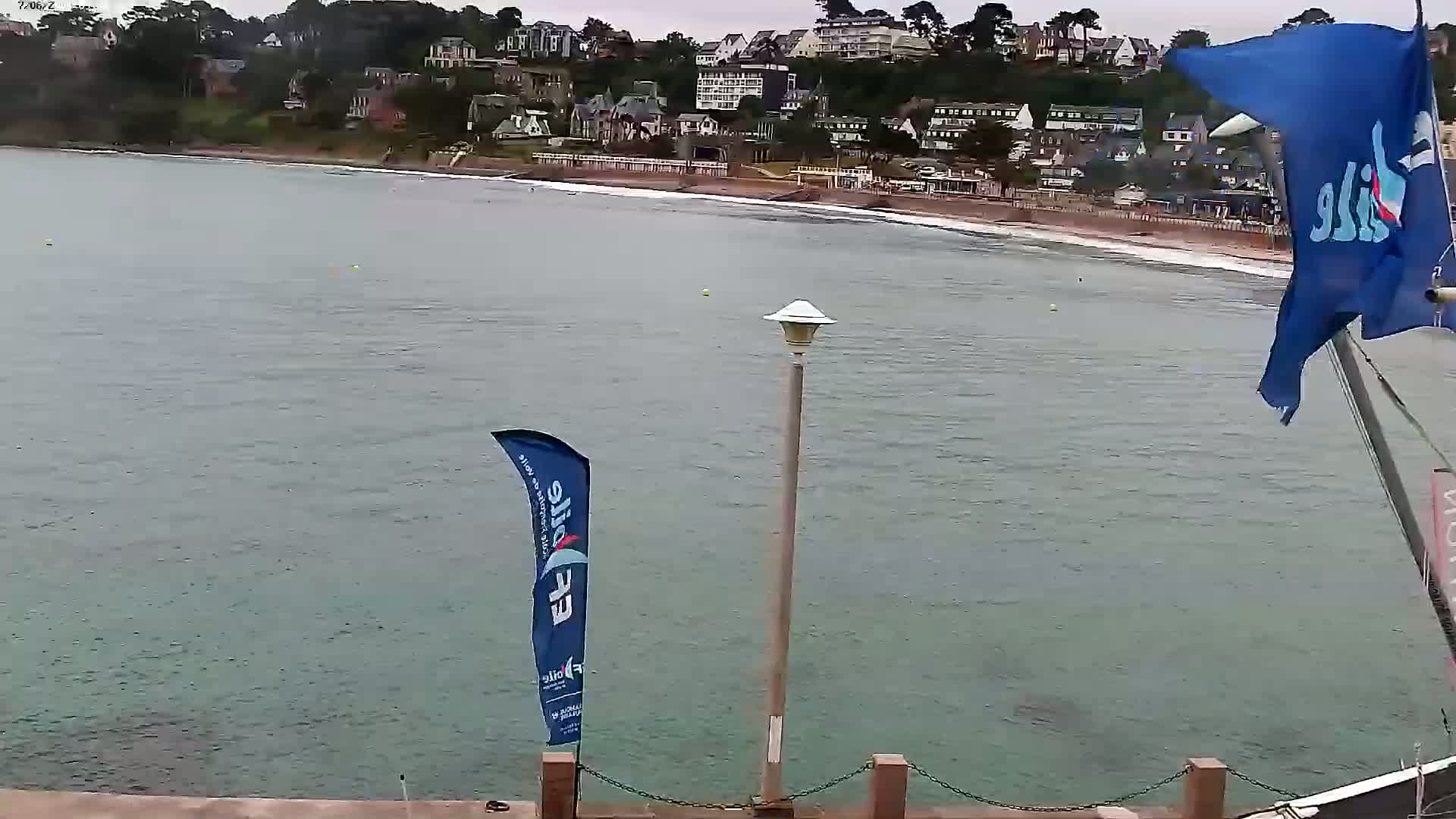 A calm body of water laps a sandy beach in front of a hillside town on an overcast day.