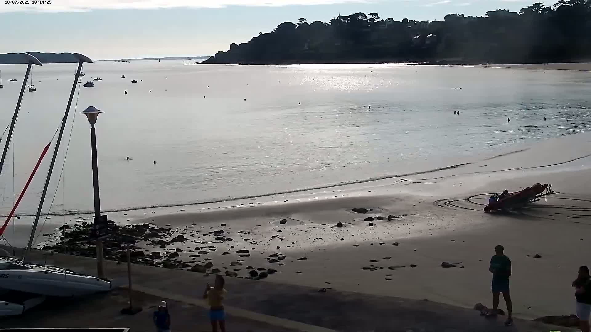 A sandy beach with calm water and several people, including some swimmers and people near a small boat on the shore, under a mostly clear sky.