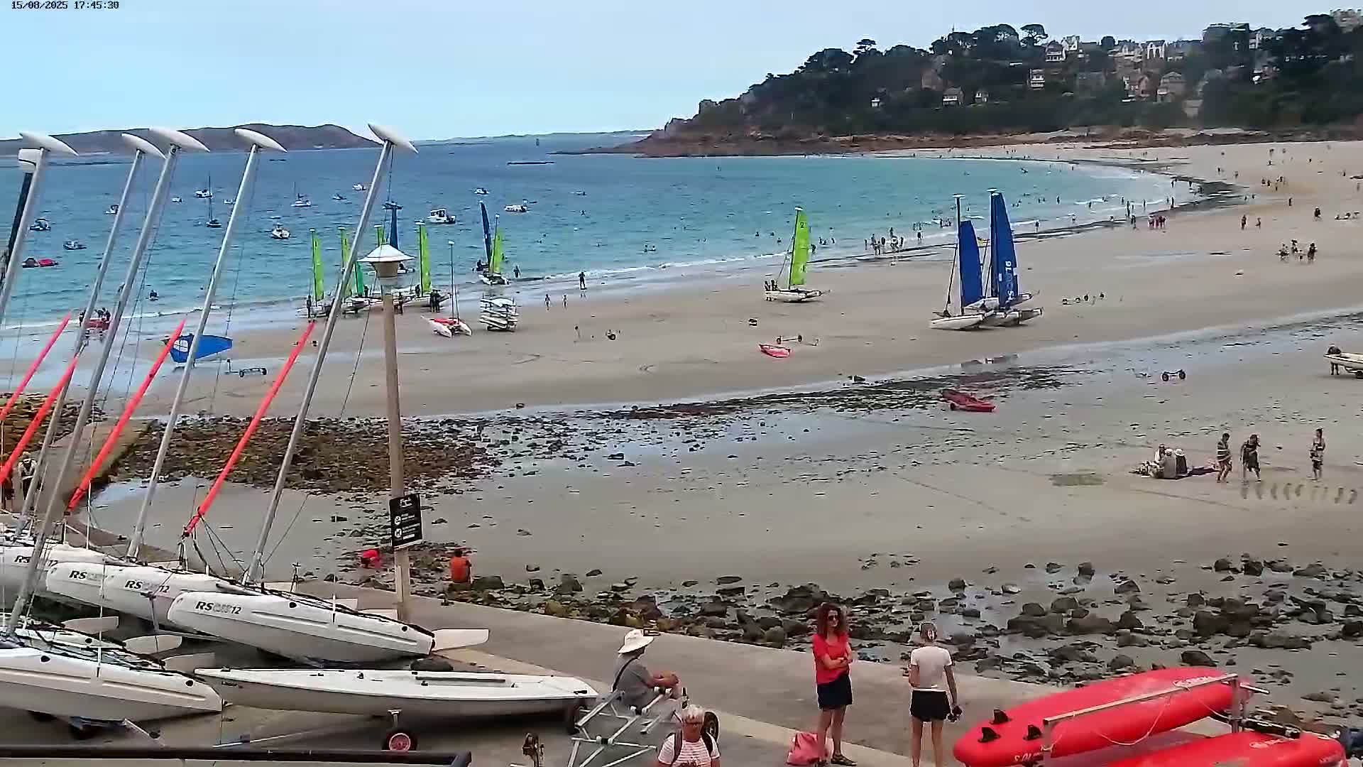 A sandy beach with calm water and several people, including some swimmers and people near a small boat on the shore, under a mostly clear sky.