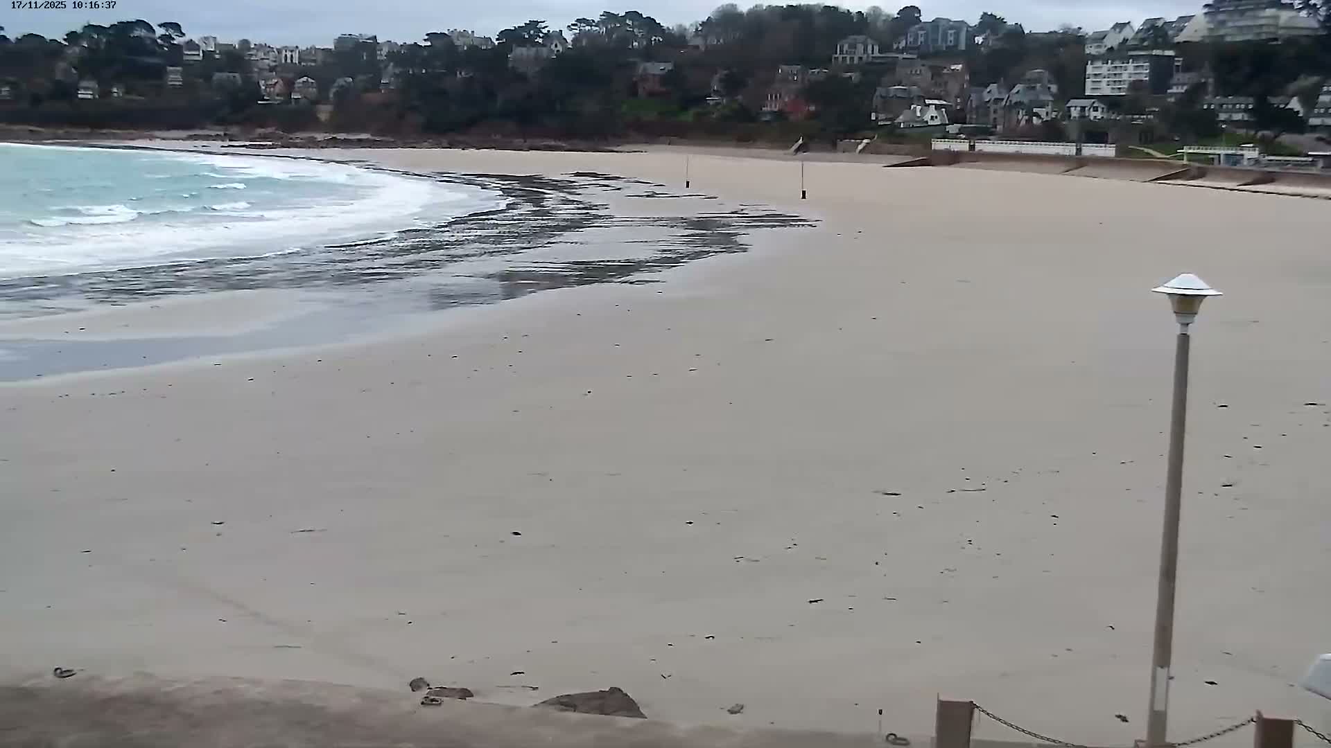 A wide sandy beach with pale blue waves breaking on the left and a town-covered hillside in the background is depicted under an overcast sky, featuring a lamppost in the right foreground.