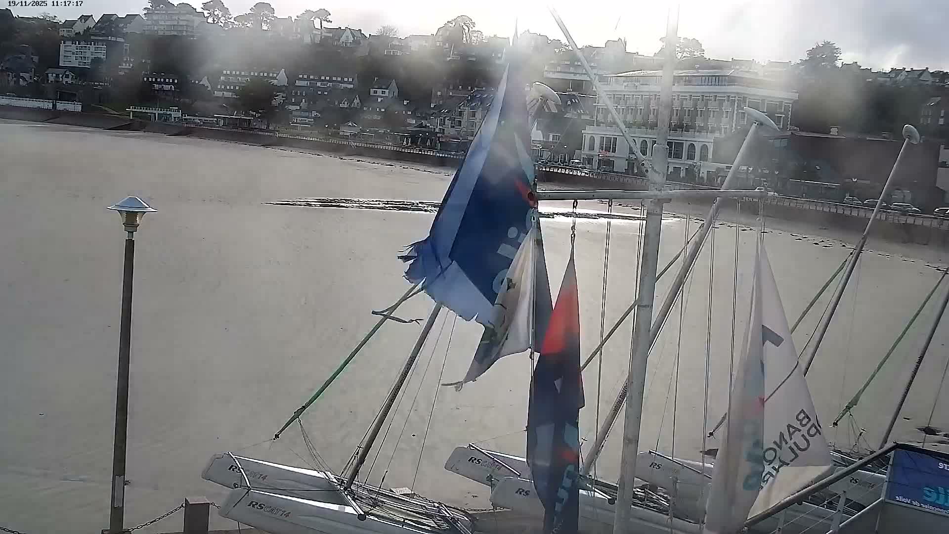 Several small sailboats and catamarans are docked on a wide sandy beach at low tide, with a town sprawling up a hillside in the background, all bathed in the bright, hazy light of a sun-glared sky.