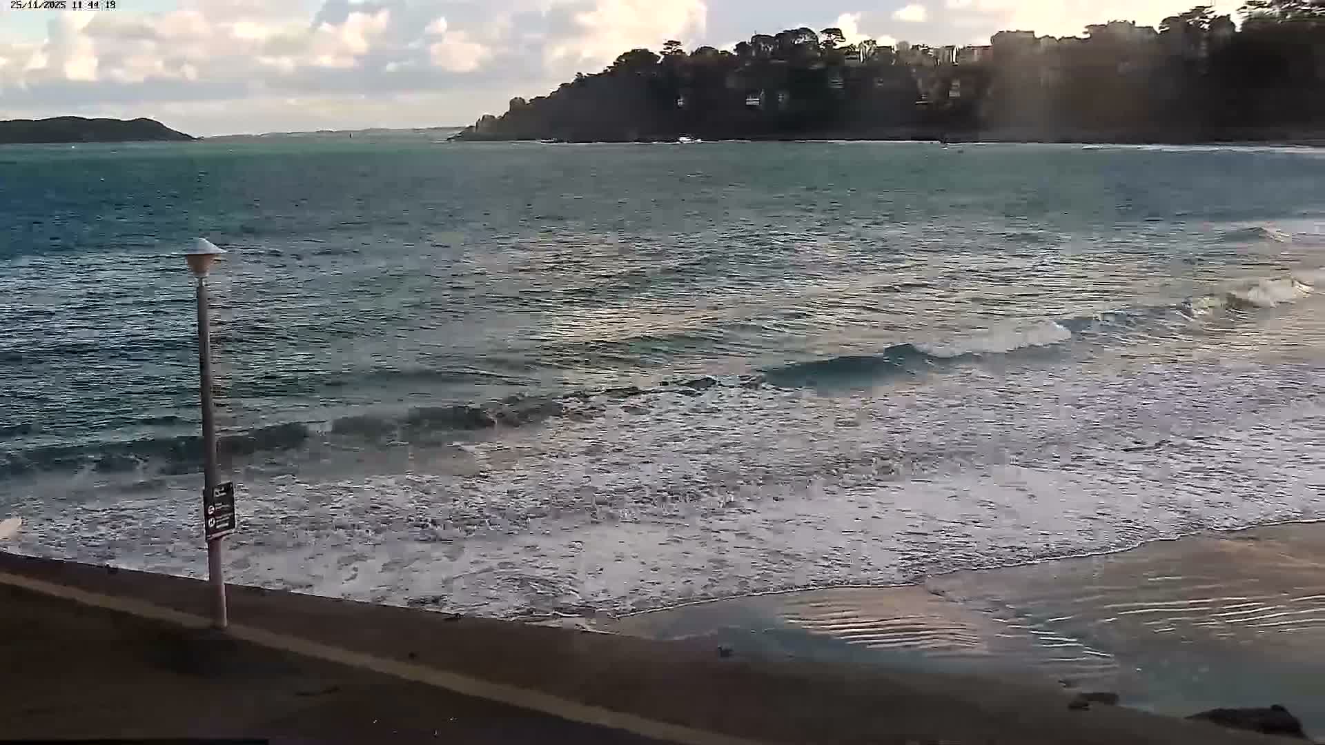 Gentle waves roll onto a sandy beach from a blue-green sea, bordered by a dark promenade with a lamppost, all beneath a partly cloudy sky with some sun peeking through, revealing a tree-covered headland in the distance.