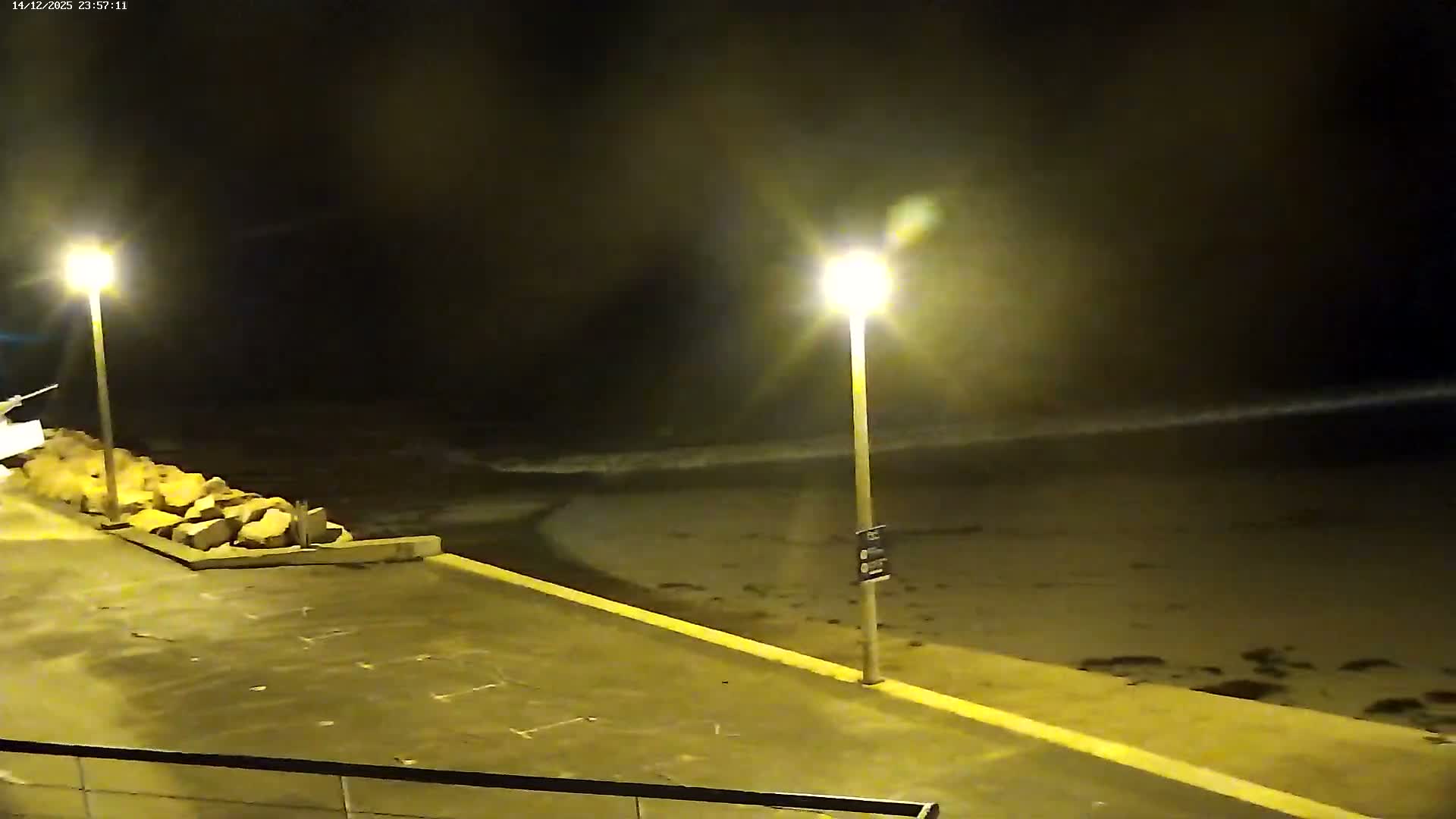 Gentle waves roll onto a sandy beach from a blue-green sea, bordered by a dark promenade with a lamppost, all beneath a partly cloudy sky with some sun peeking through, revealing a tree-covered headland in the distance.