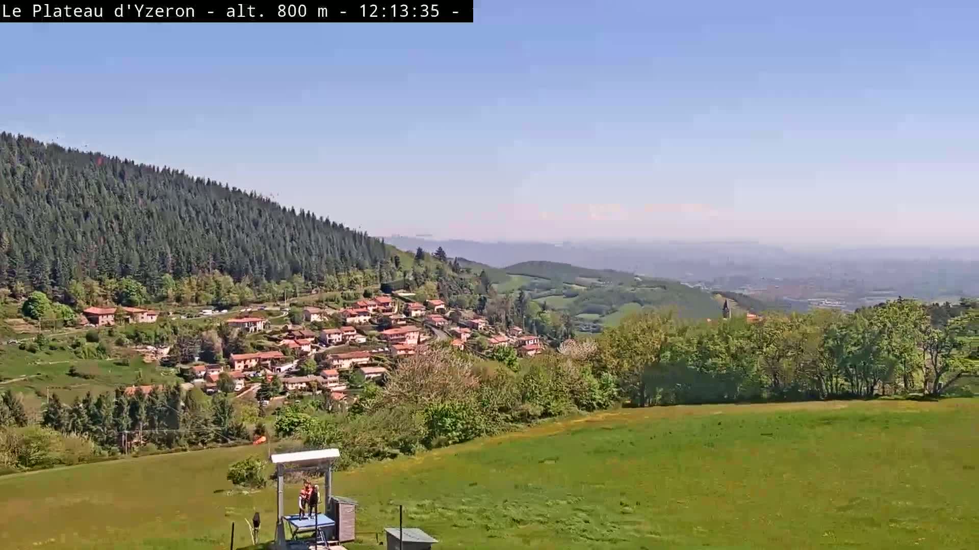 A small village nestled in a valley, surrounded by green hills and forests under a clear blue sky.
