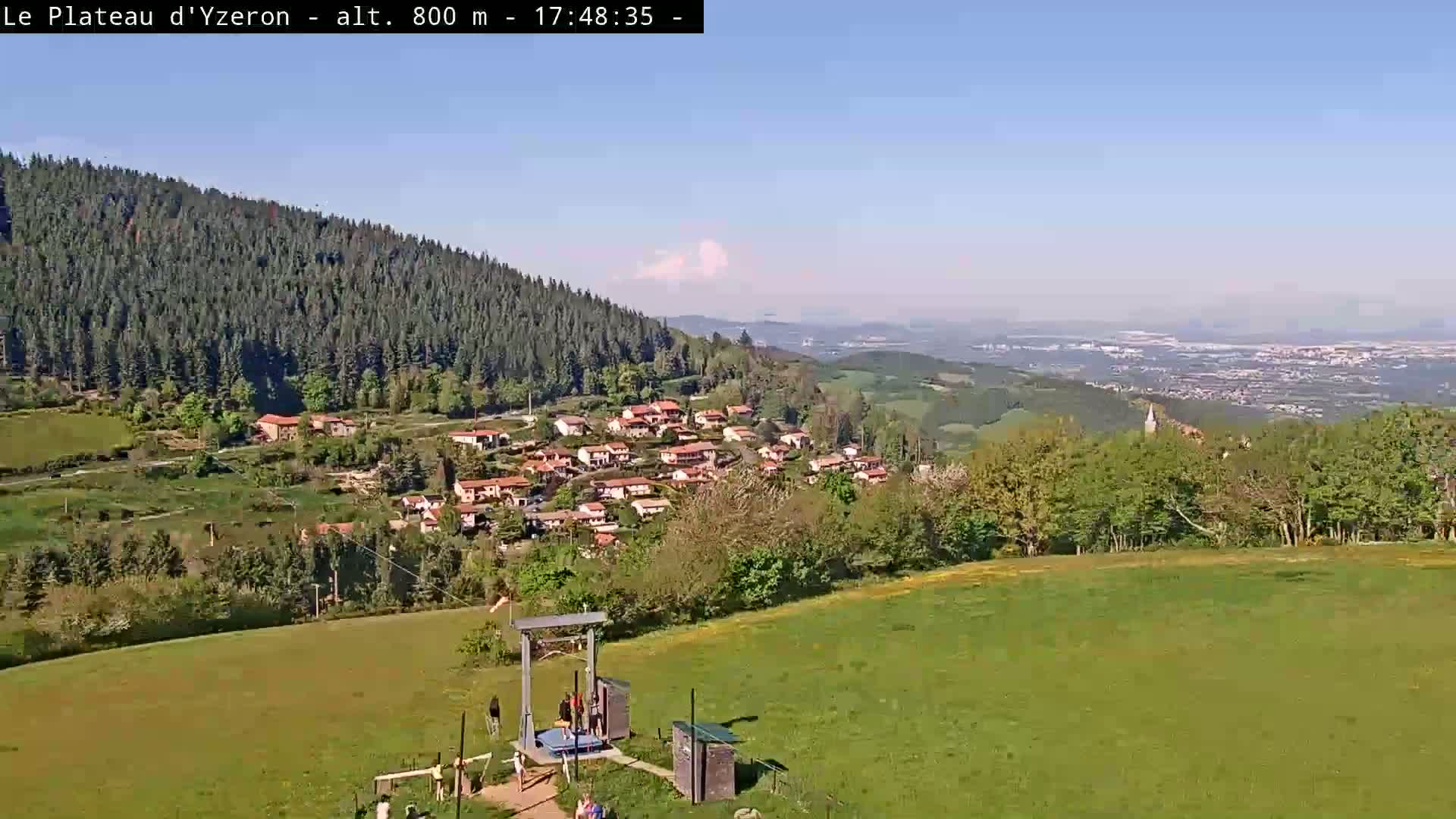 A sunny day reveals a village nestled in a valley below a verdant hillside, with a distant cityscape visible in the background.