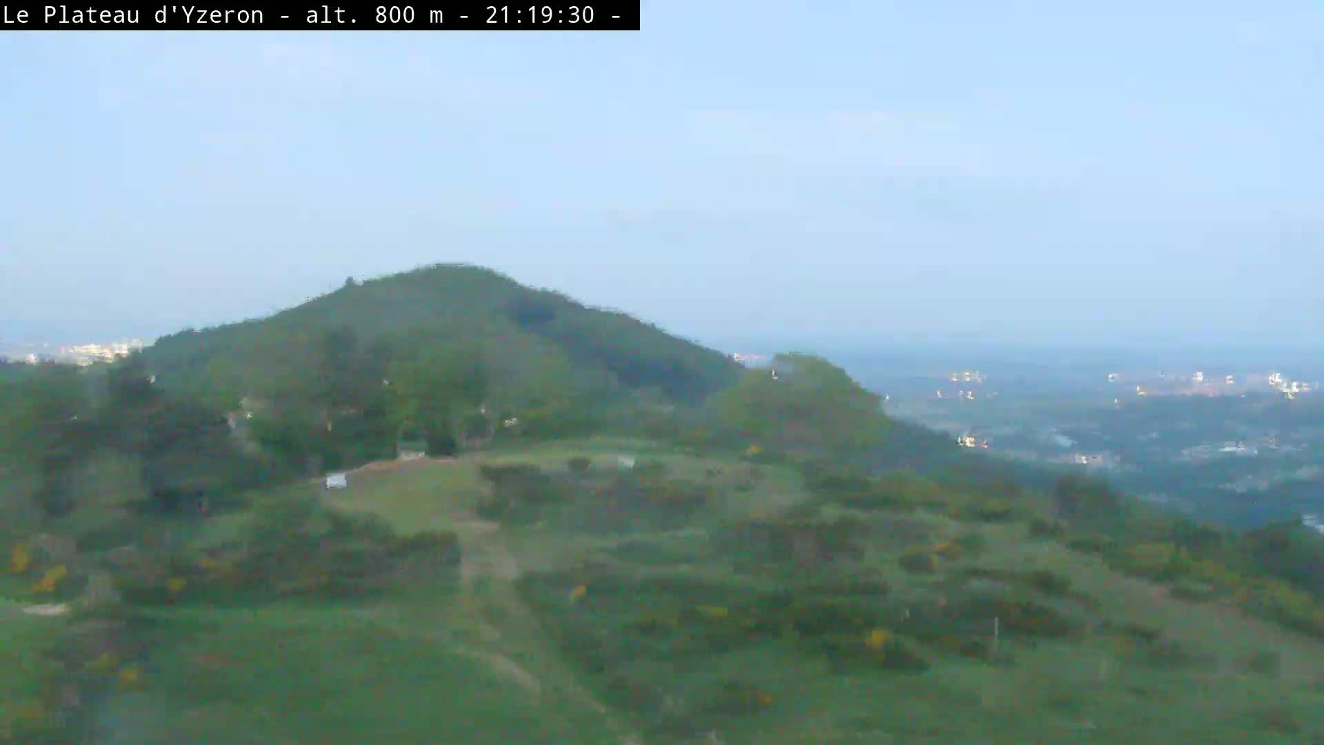 A hazy, twilight view shows a gently rolling green hilltop overlooking a distant city at night.