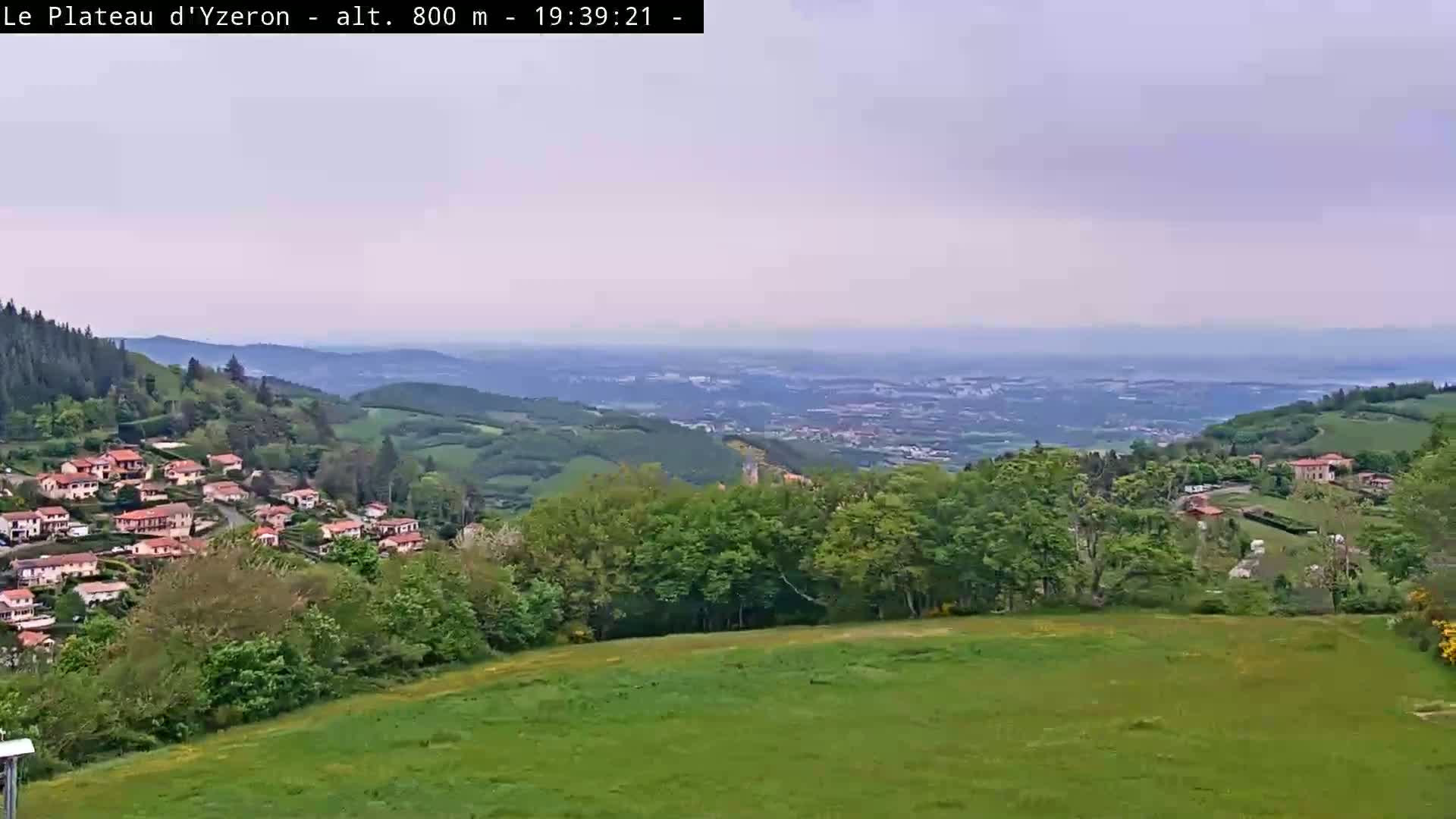 A hazy, overcast view shows a village nestled in a verdant valley, overlooking a sprawling city in the distance.
