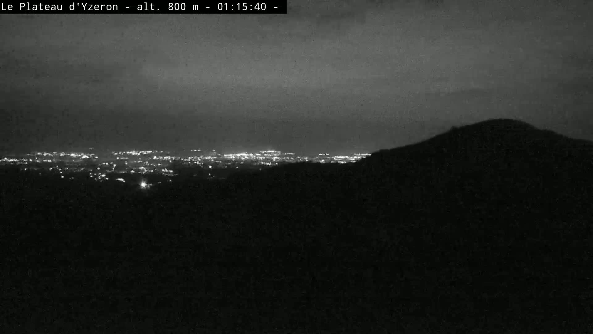 A nighttime view shows a distant city's lights spread across a valley, partially obscured by a dark hill or mountain in the foreground under a dark, cloudy sky.