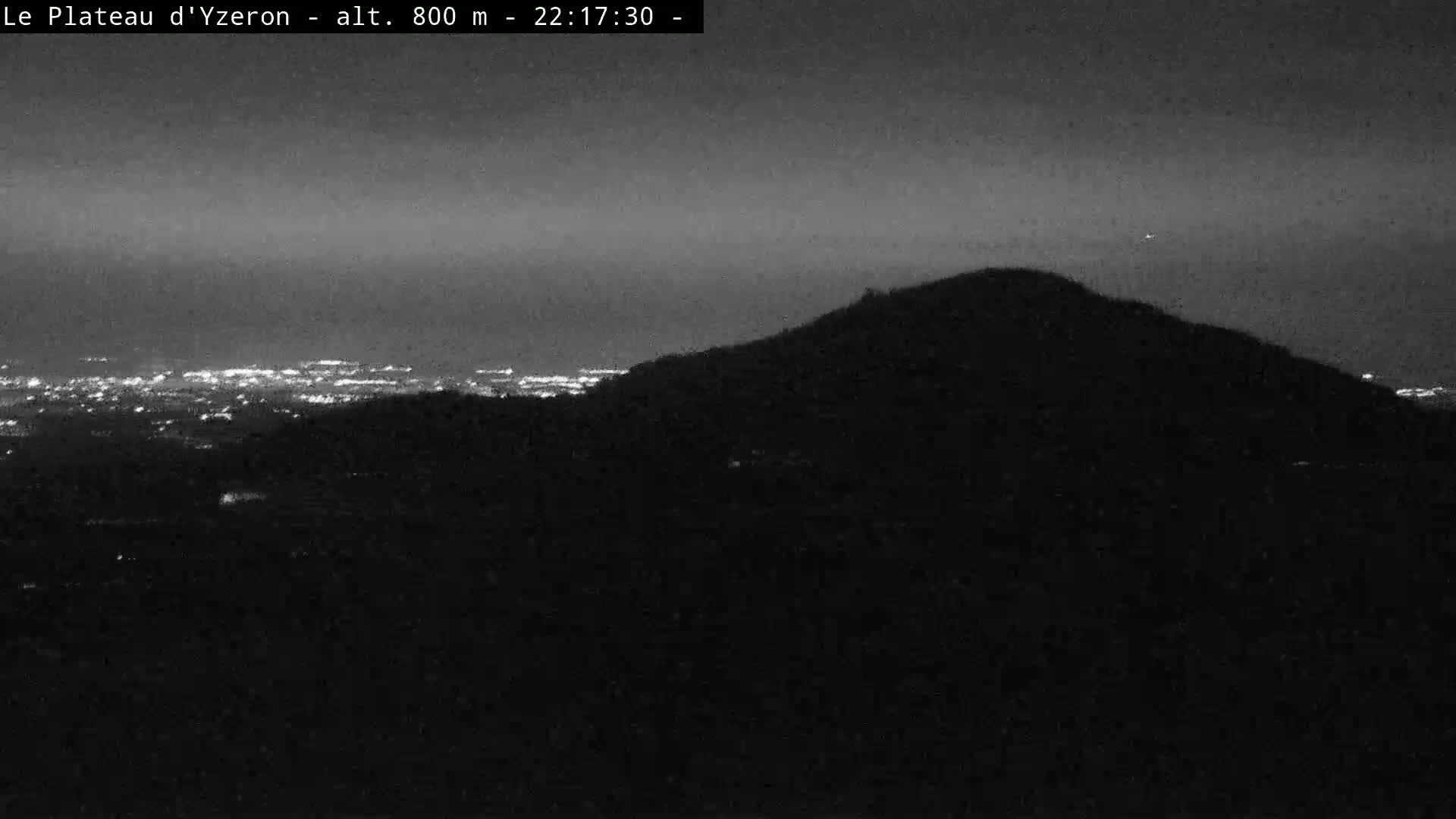 A dark, nighttime view shows a silhouetted hill overlooking a distant city illuminated by lights under a clear sky.