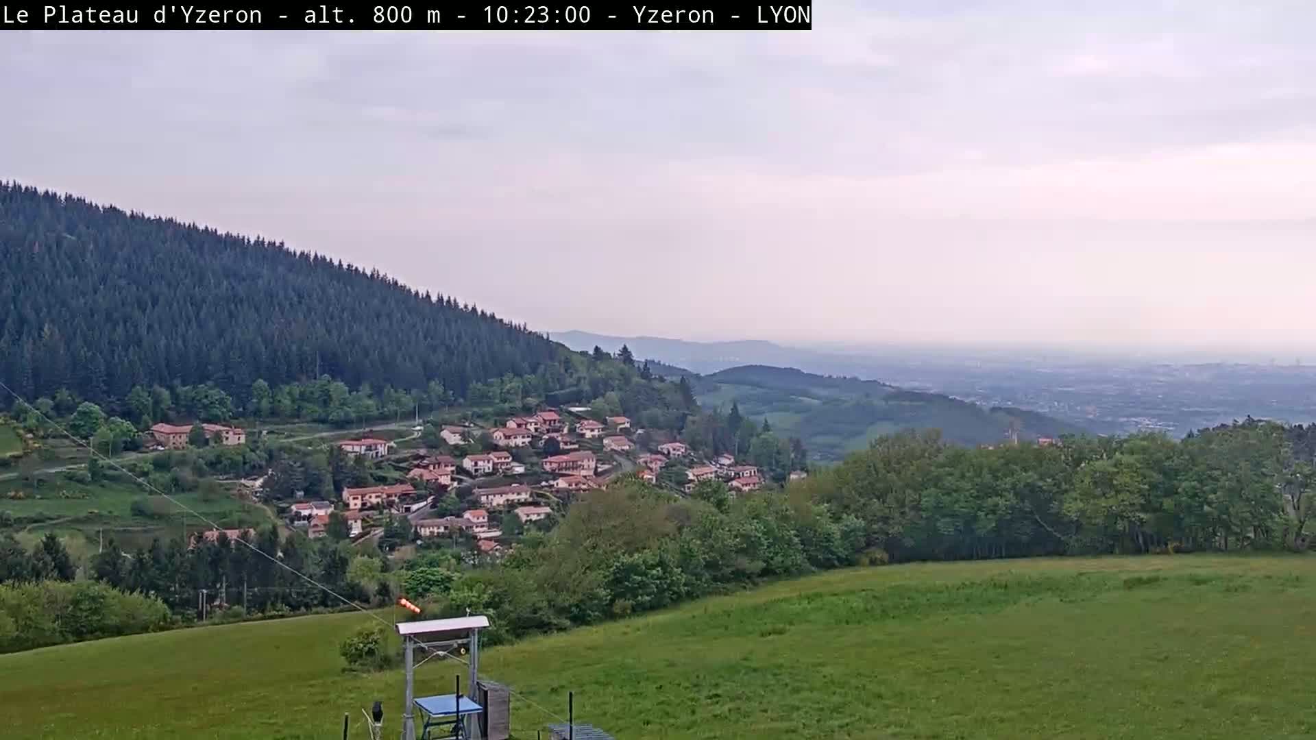 A village nestled in a valley below a forested hillside, with a hazy view of a distant city under a partly cloudy sky.