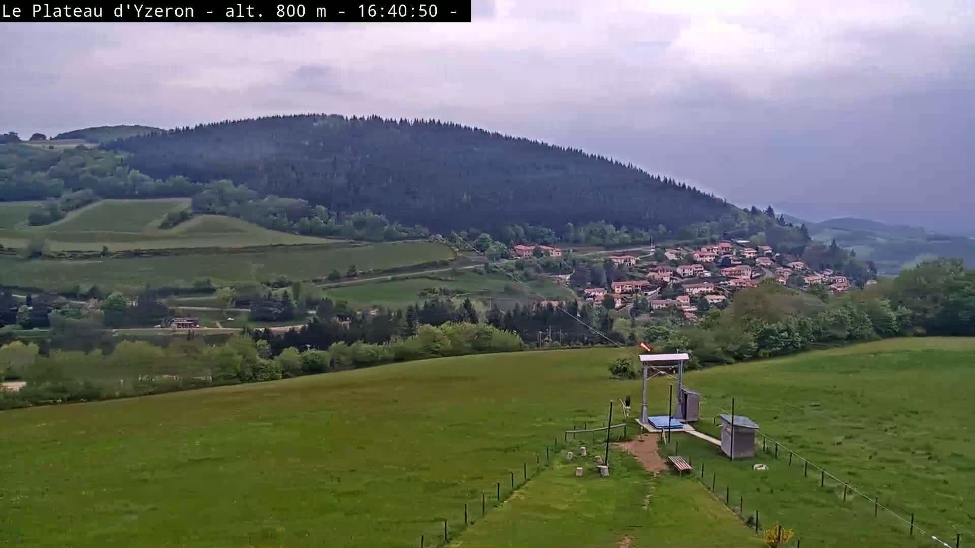 A verdant hillside with a small village nestled in the distance under a cloudy sky, features a zipline launch in the foreground.