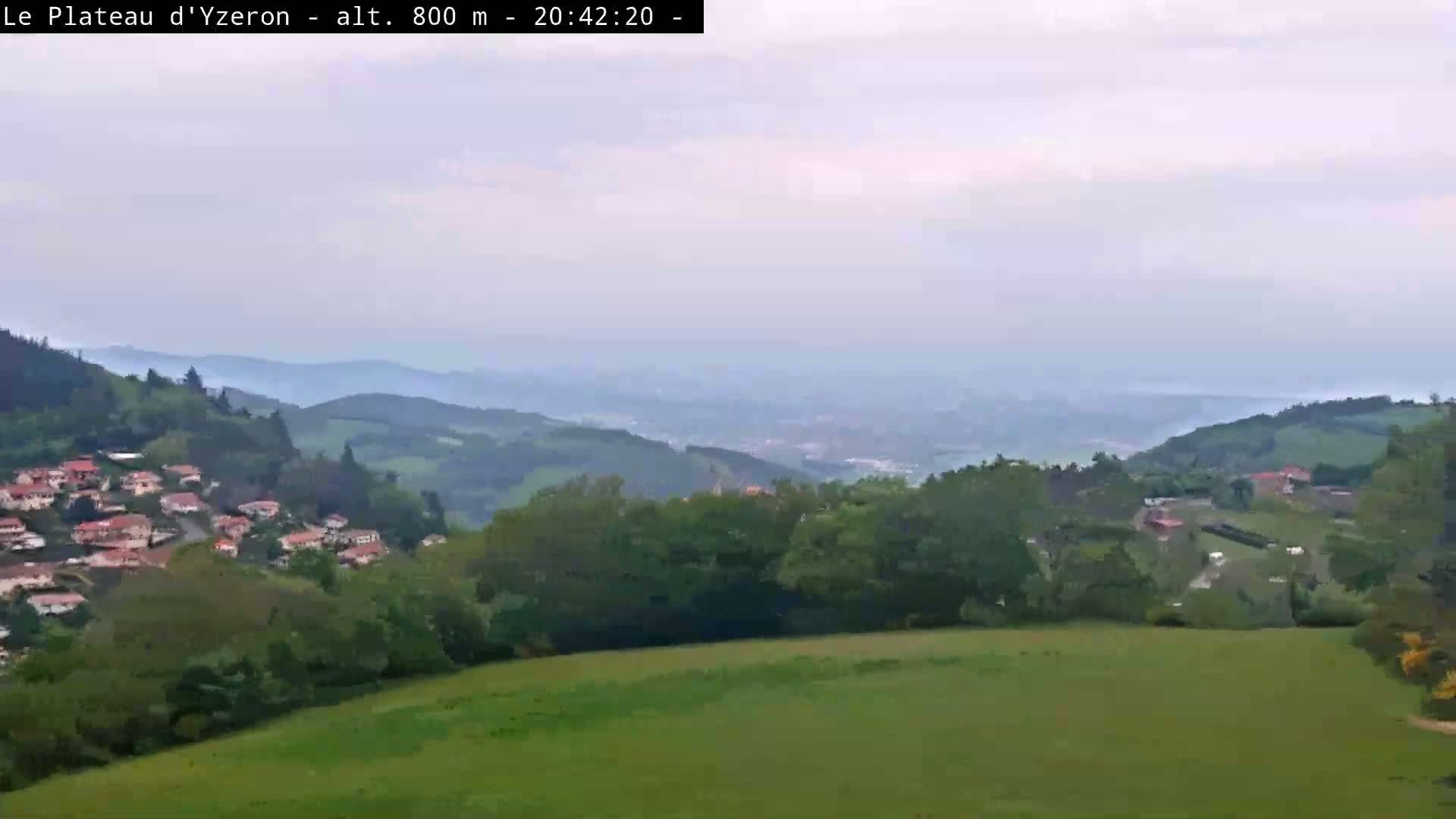 A hazy view shows rolling green hills and a small village nestled amongst them, overlooking a distant city under an overcast sky.