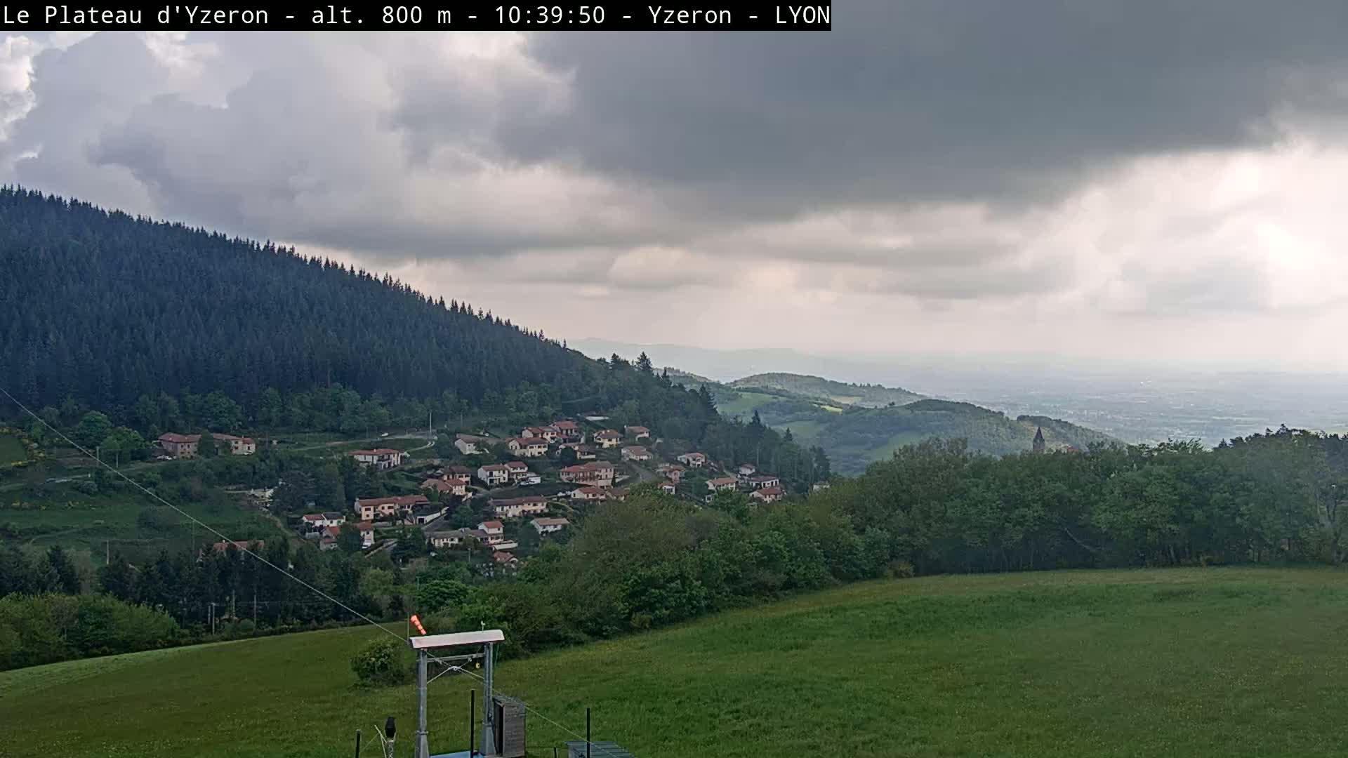 A village nestled in a hillside valley under an overcast sky, with a vast expanse of green fields and distant mountains visible in the background.