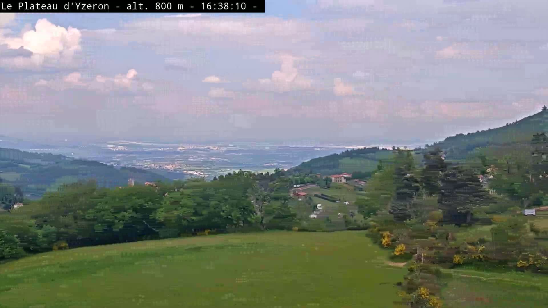 A partly cloudy sky overlooks a verdant valley dotted with trees and buildings, extending to a distant hazy plain.