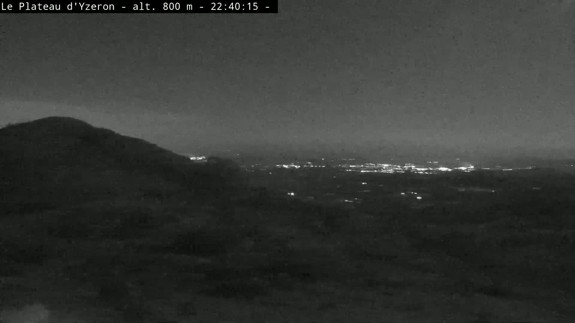 A nighttime, low-light view shows a hill overlooking a distant city illuminated by lights under a dark sky.