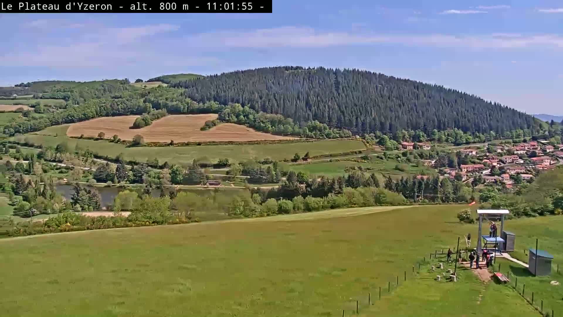 A sunny view of a green valley with a village, a lake, and a forested hillside, featuring a zip line at the foreground.