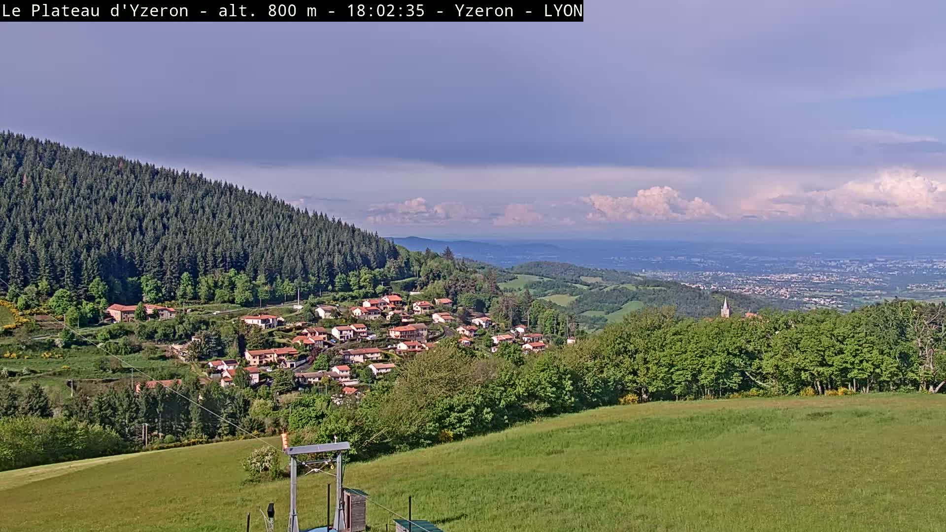 A small village nestled in a hillside, surrounded by lush green fields and forests, overlooks a distant city under a partly cloudy sky.