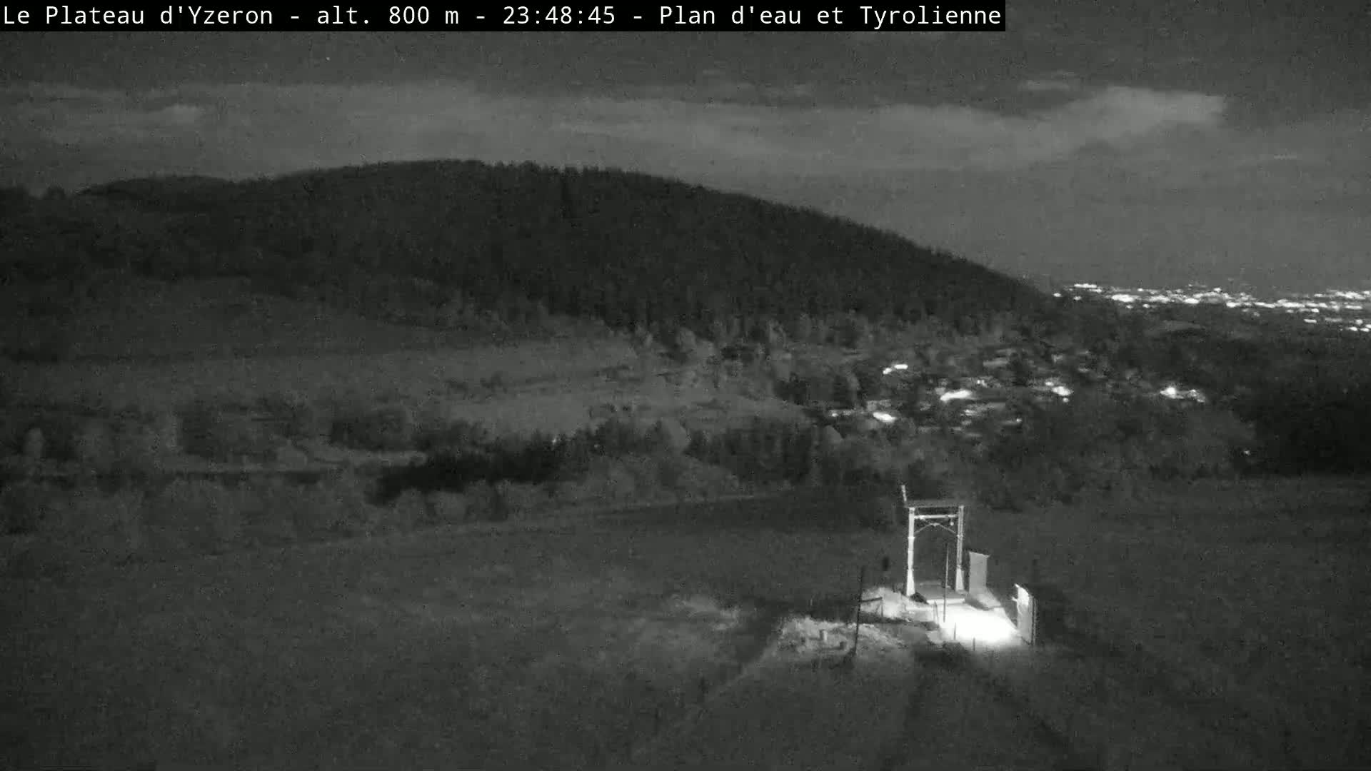 A nighttime, low-light view shows a hilly landscape with a small structure in the foreground under a mostly clear sky.