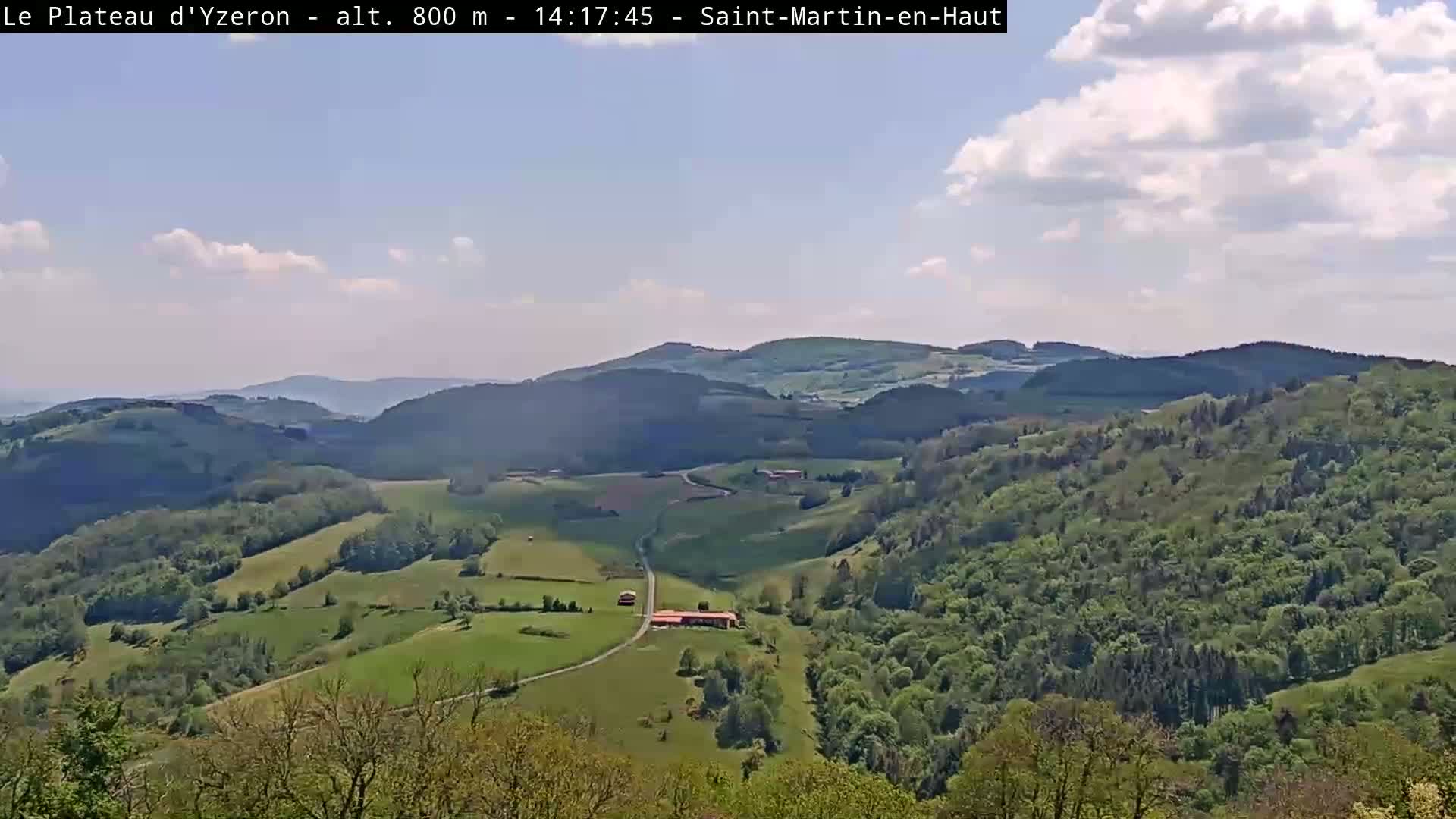 A partly cloudy day reveals a panoramic view of rolling green hills and valleys dotted with trees and a few buildings.