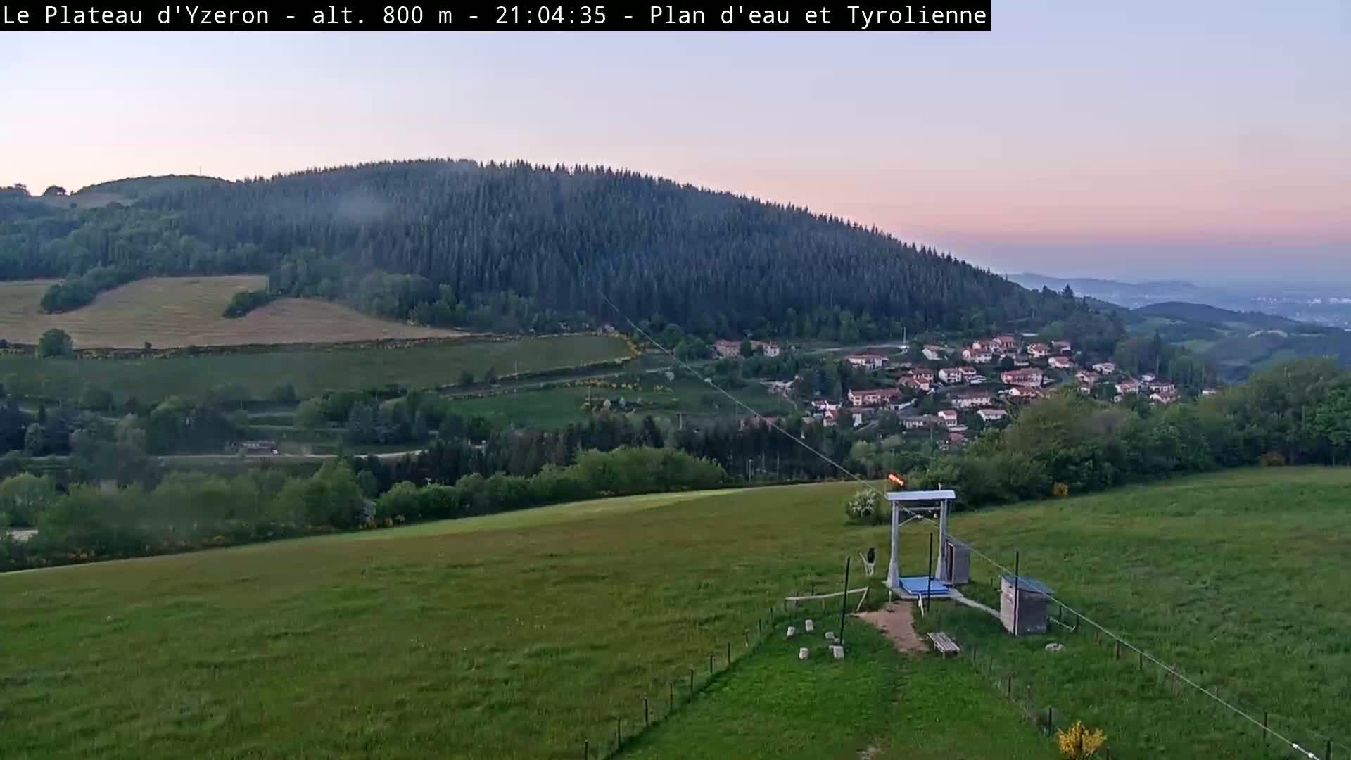 A grassy field with a small structure and a cable stretches towards a village nestled in a valley below a large forested hill, all under a twilight sky.
