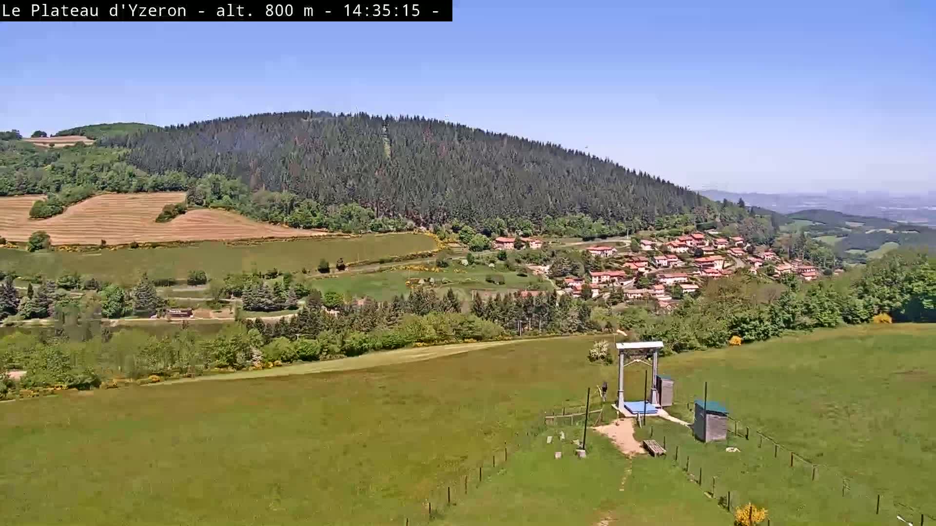 A sunny view shows a green valley with a small village nestled amongst the trees, a ski lift in the foreground, and rolling hills extending to the horizon under a clear blue sky.
