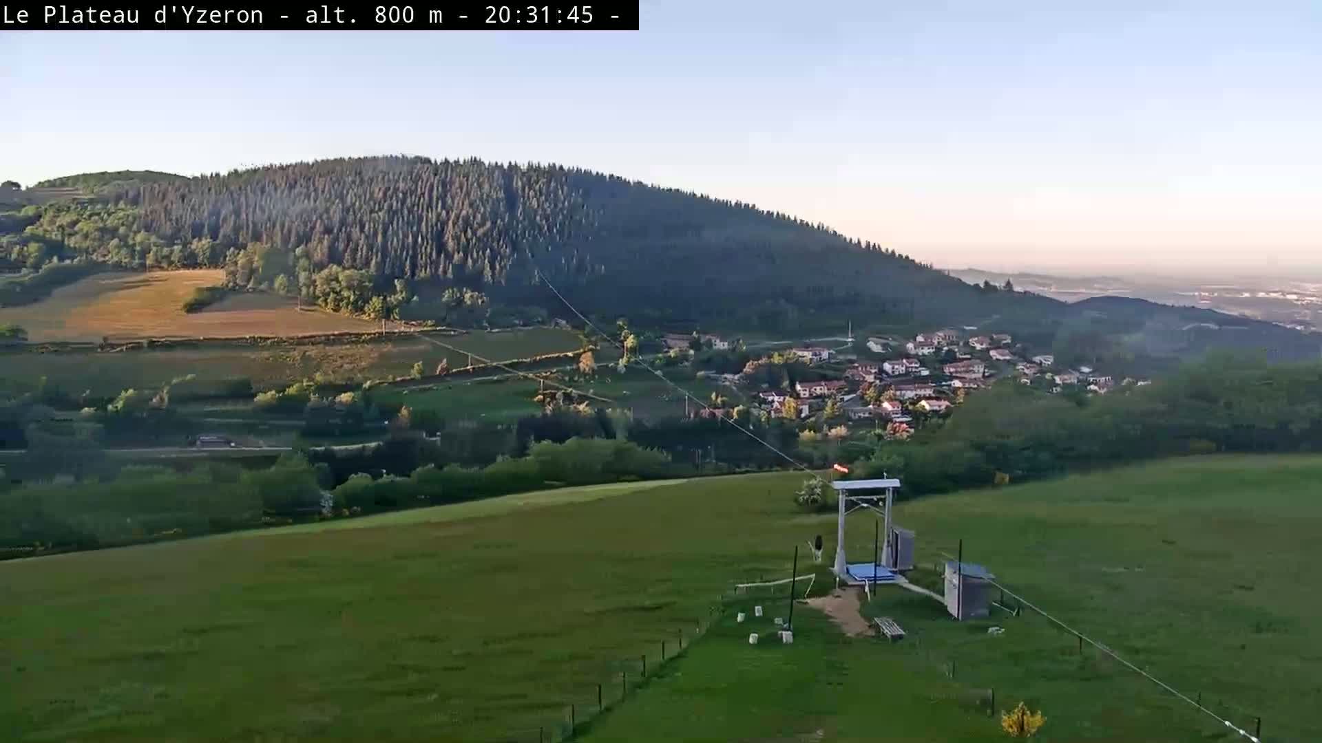 A high-angle view shows a grassy field with a zipline apparatus in the foreground, overlooking a village nestled in a valley beneath a forested hill, all under a clear, bright sky.