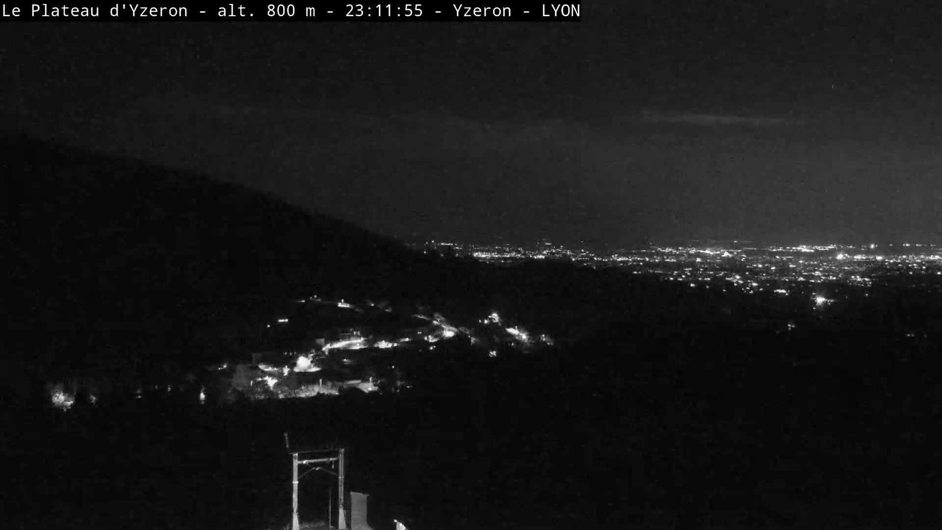 A nighttime, long-range view shows a city's lights in the distance, with a darker foreground showing a small town and hills.